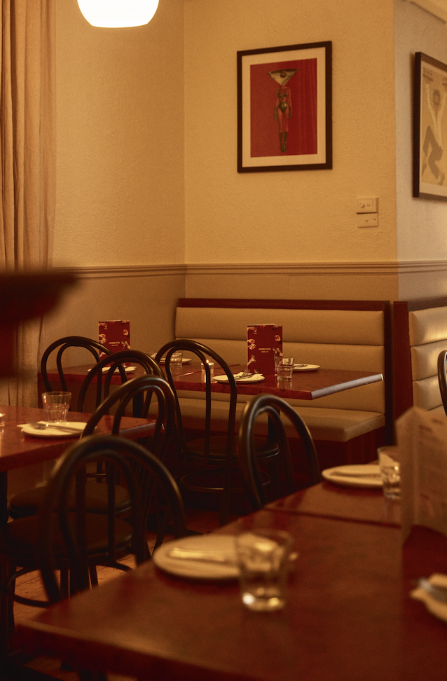 An empty restaurant with a warm, dimly lit ambiance. There are wooden tables set with plates, glasses, and napkins. The seating includes black chairs and a white banquette with red accents. A framed painting hangs on the wall above the banquette, and a ceiling light fixture provides soft lighting.