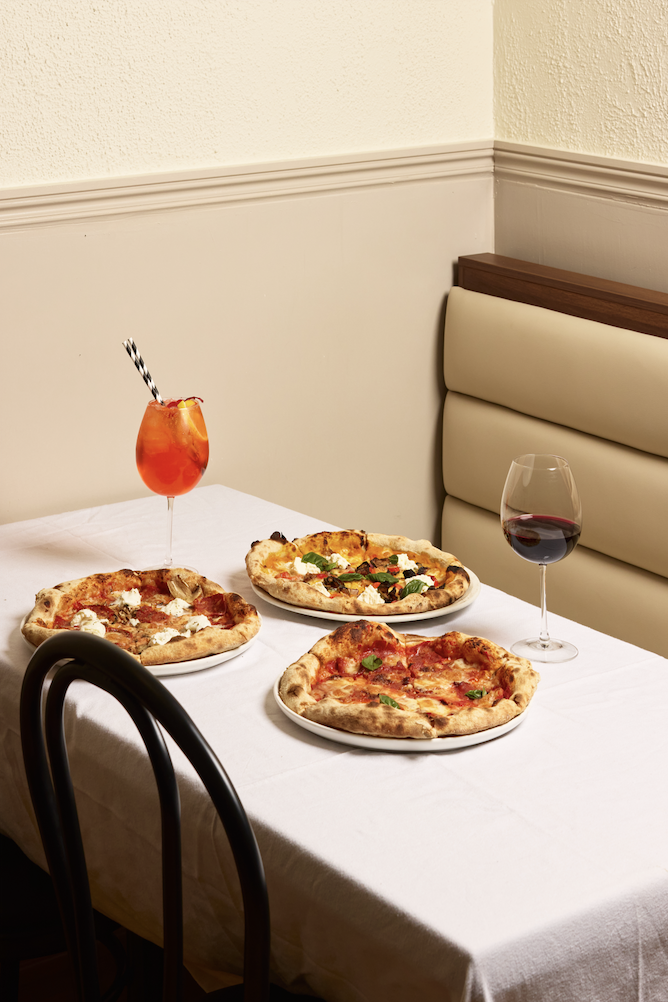 Table with three pizzas, a glass of red wine, and a cocktail in a restaurant setting