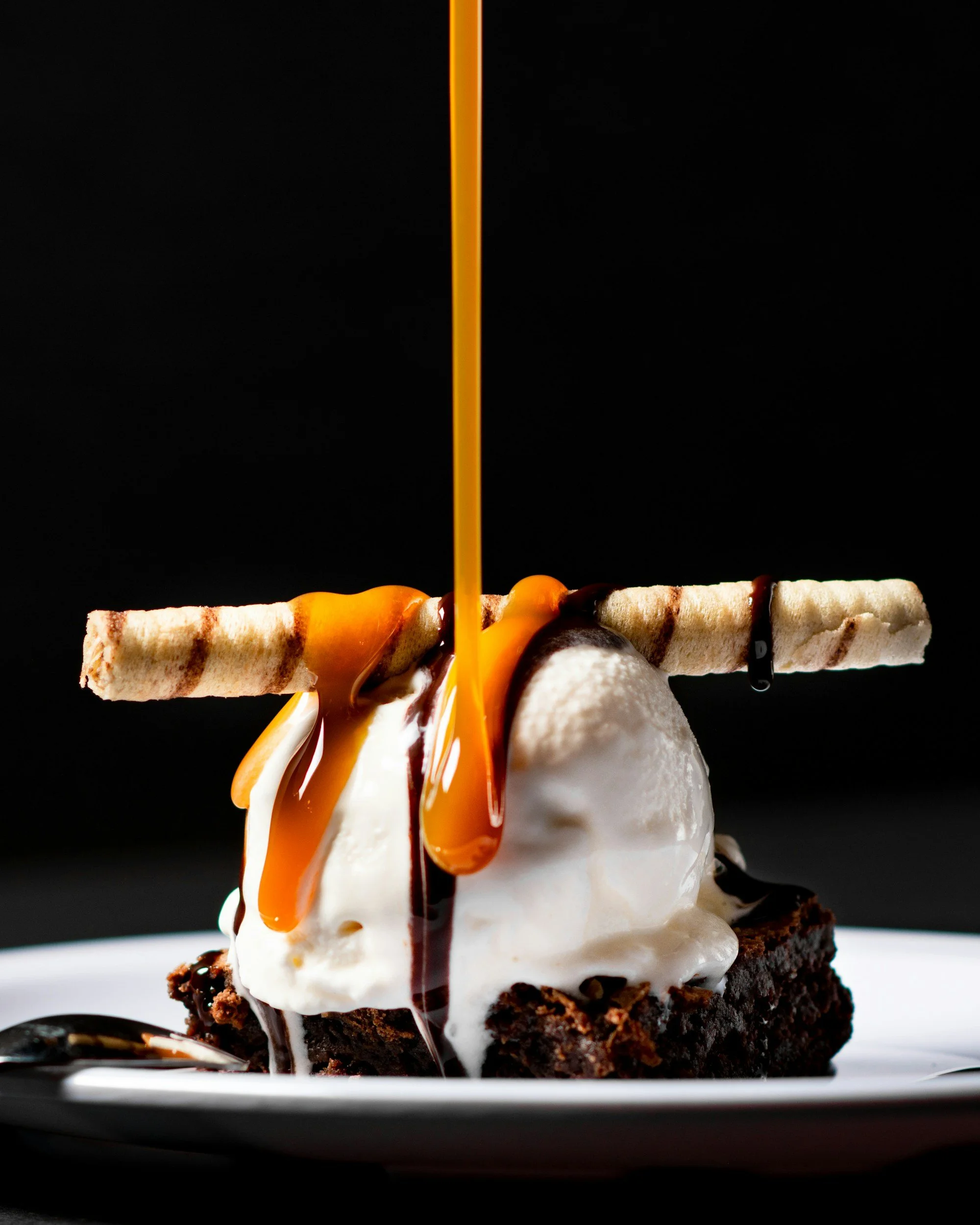 Chocolate brownie with a scoop of vanilla ice cream, a rolled wafer cookie, caramel and chocolate syrup being poured over the top placed on a white plate against a black background.