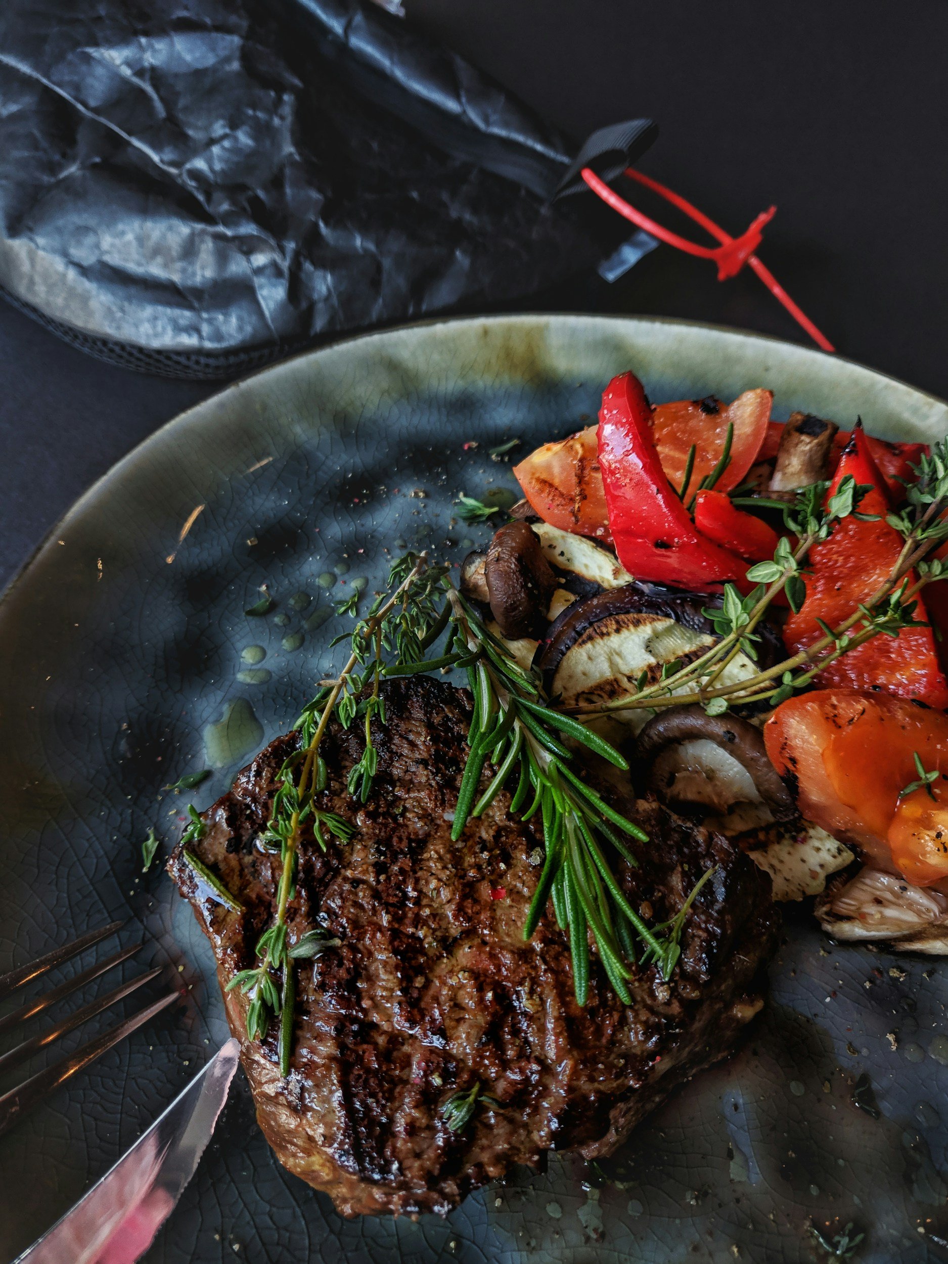 A cooked steak topped with rosemary on a black plate, with grilled vegetables like red bell peppers, mushrooms, and zucchini nearby.