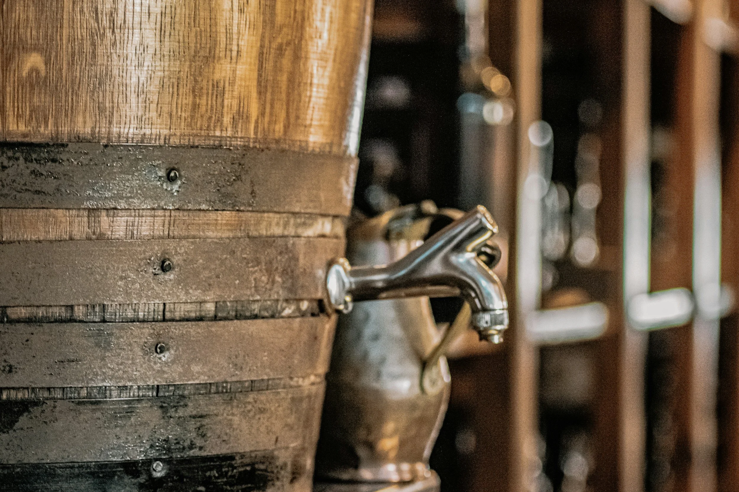 Close-up of a vintage metal water tap attached to a wooden barrel or container, with a blurred background of wooden shelves.