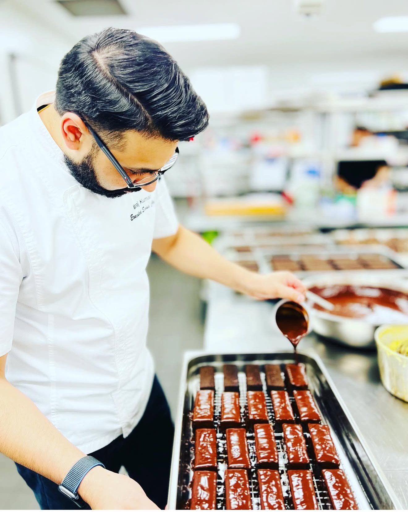 A chef pours chocolate glaze onto rectangular pastry treats arranged on a tray in a commercial kitchen.