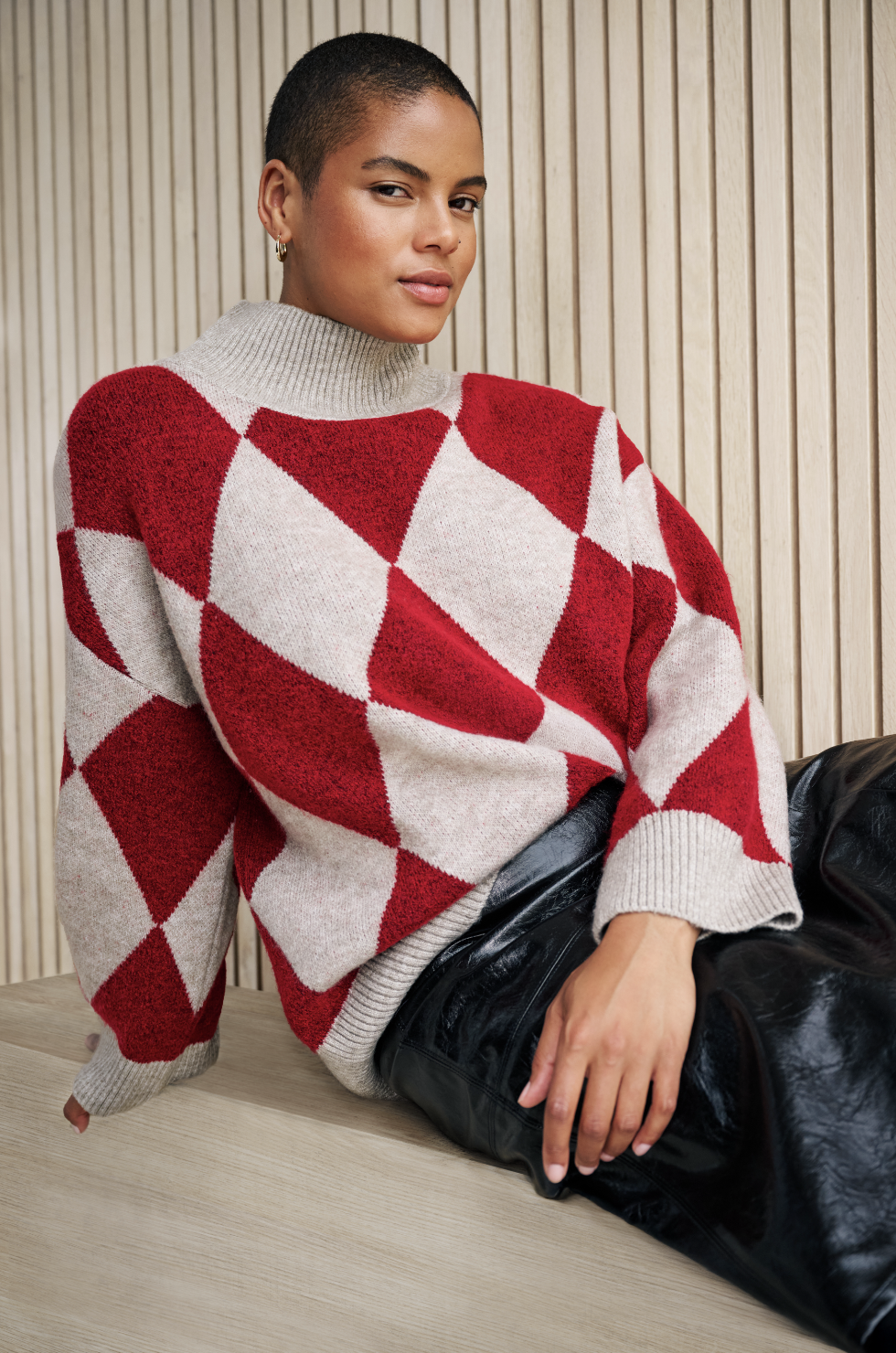A woman with short hair wearing a beige and red argyle sweater sitting on a wooden table against a wooden paneled wall.