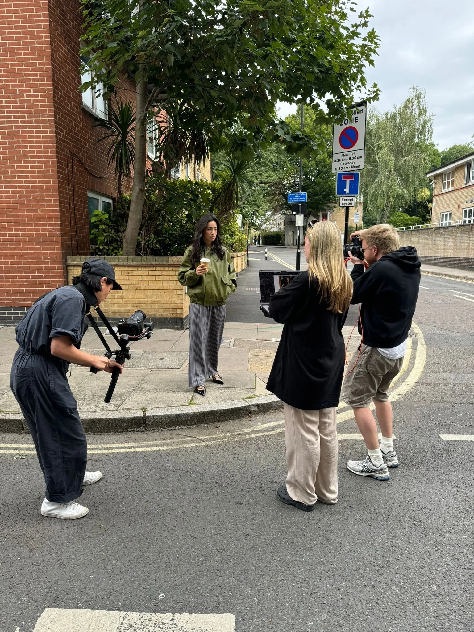 A group of four people filming a woman holding a coffee cup on a city sidewalk. The woman stands near a tree, and the crew includes a person holding a camera stabilizer, another using a camera, and a third with a laptop.