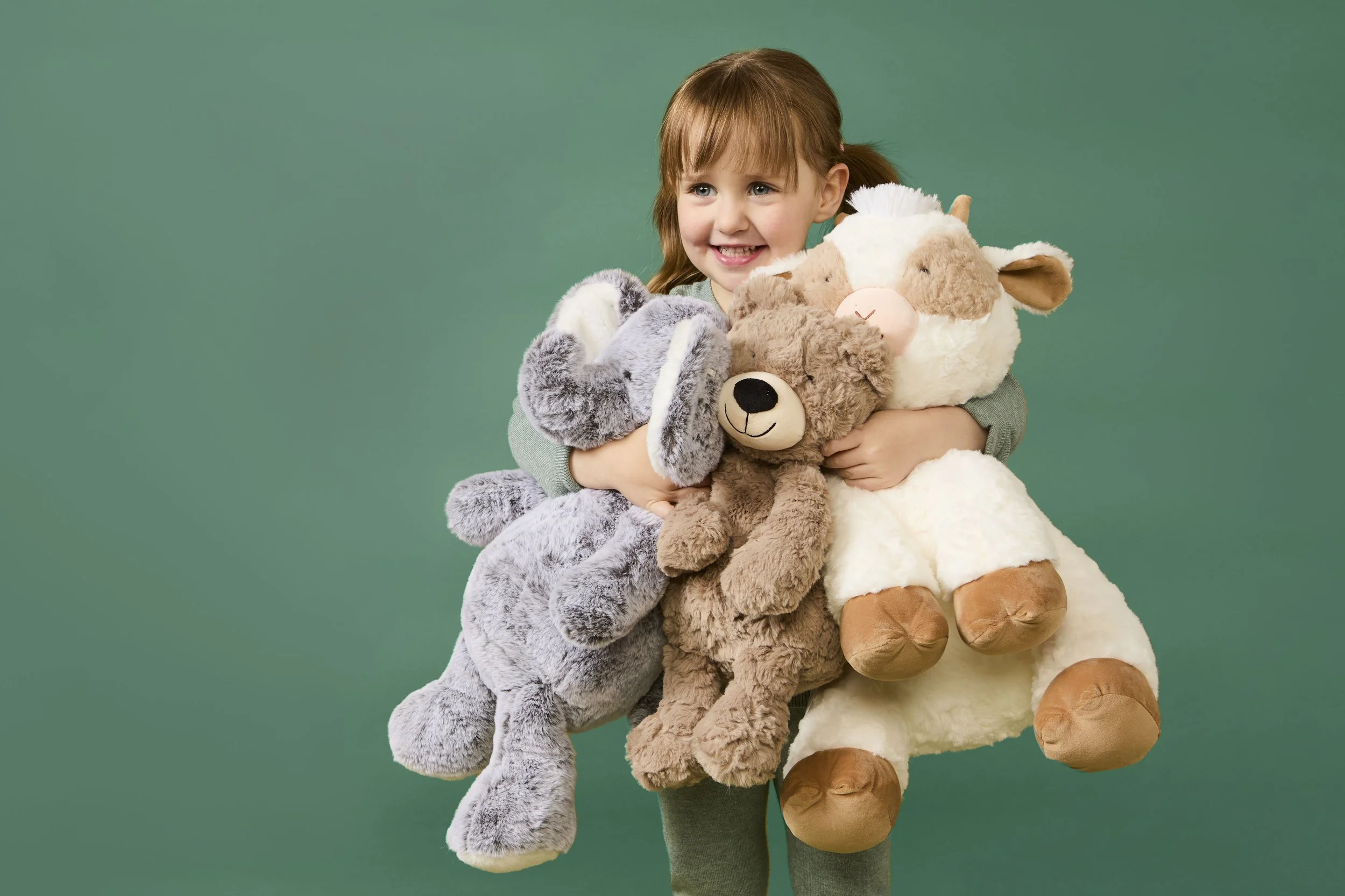 Young girl with red hair smiling and holding several large plush stuffed animals against a green background.