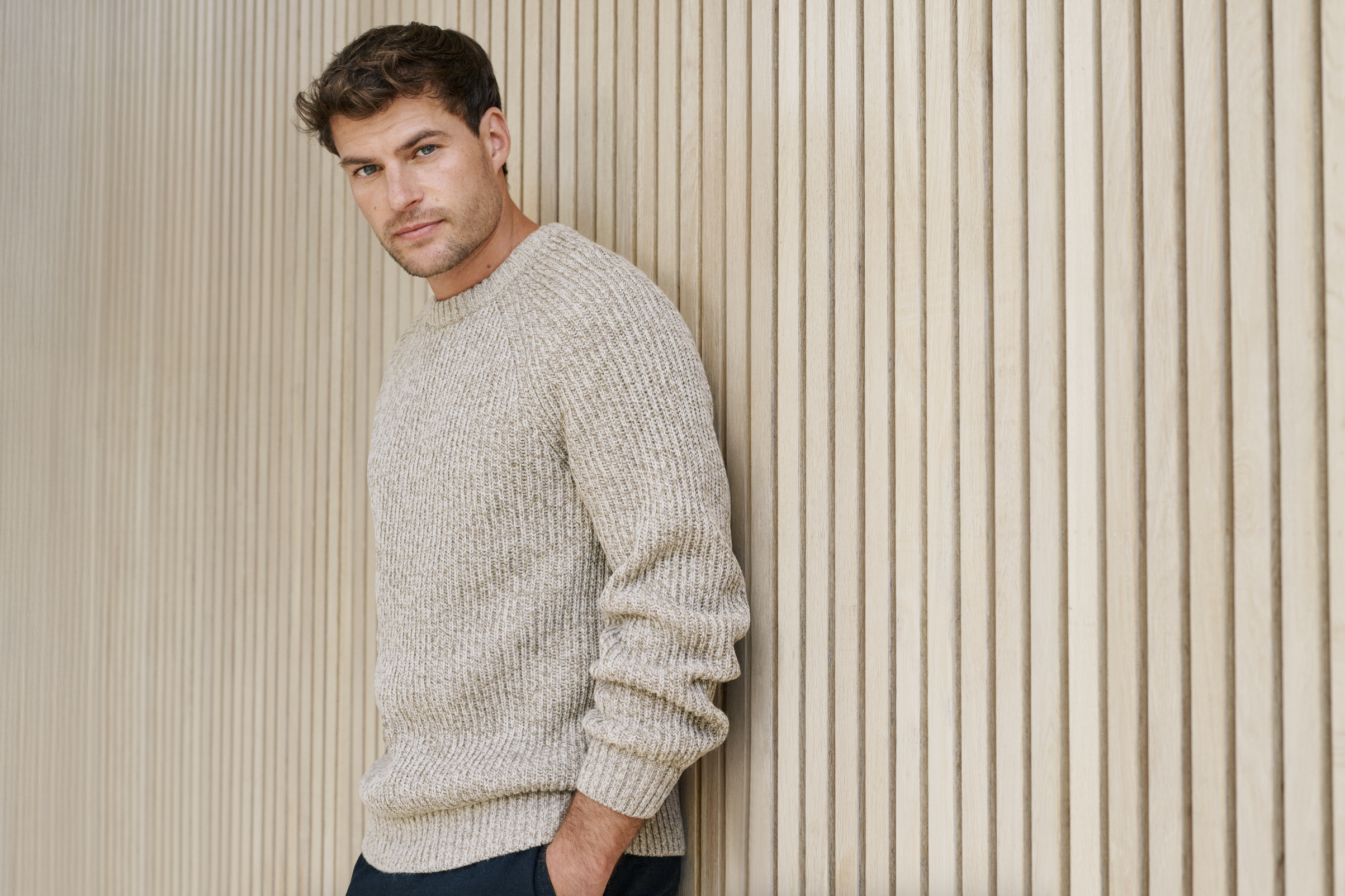 A man with brown hair and blue eyes standing against a vertically paneled beige wall, wearing a light beige knit sweater and dark pants.
