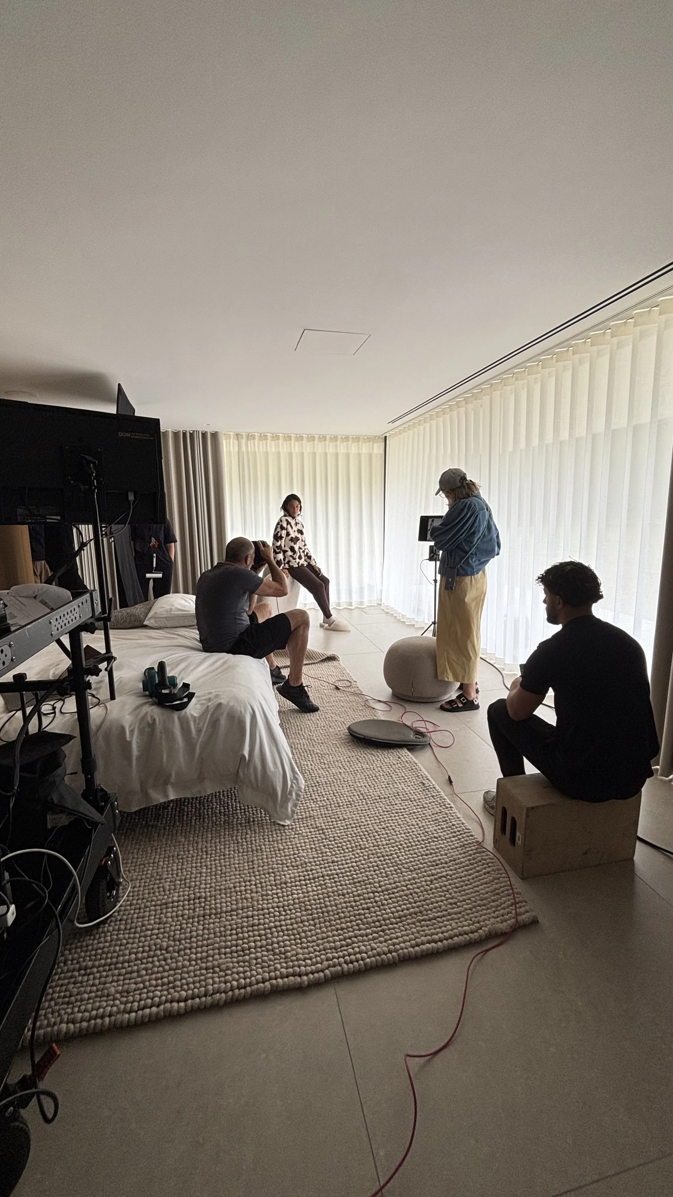Photo of a photoshoot session in a well-lit room with large curtains. A model is sitting against the curtains, and a photographer is taking pictures. Several crew members are present, including one sitting on a bed, one sitting on a box, and one operating a camera equipment.