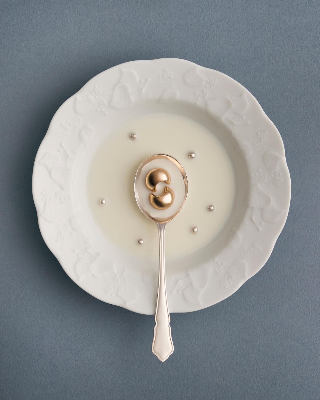 A decorative white plate with embossed floral pattern, filled with milk, with a silver spoon resting on top. The spoon holds a pair of gold earrings with a rounded design, and there are small metallic beads scattered in the milk.