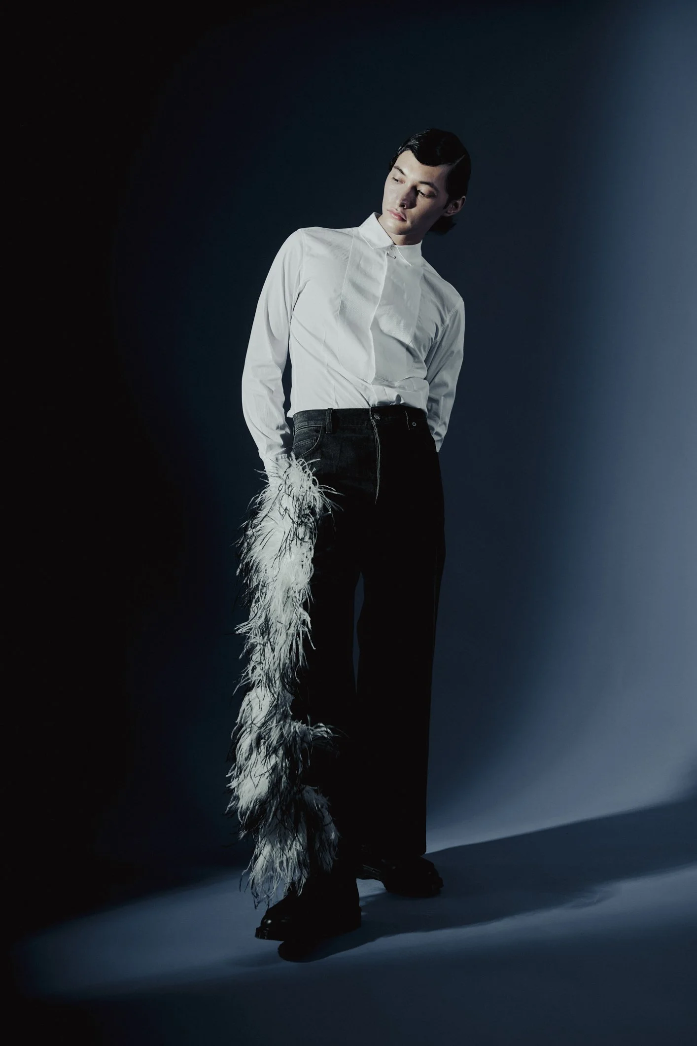 A person standing against a dark and light gradient background, wearing a white shirt with a feathered sleeve on one arm and black pants, looking downward.