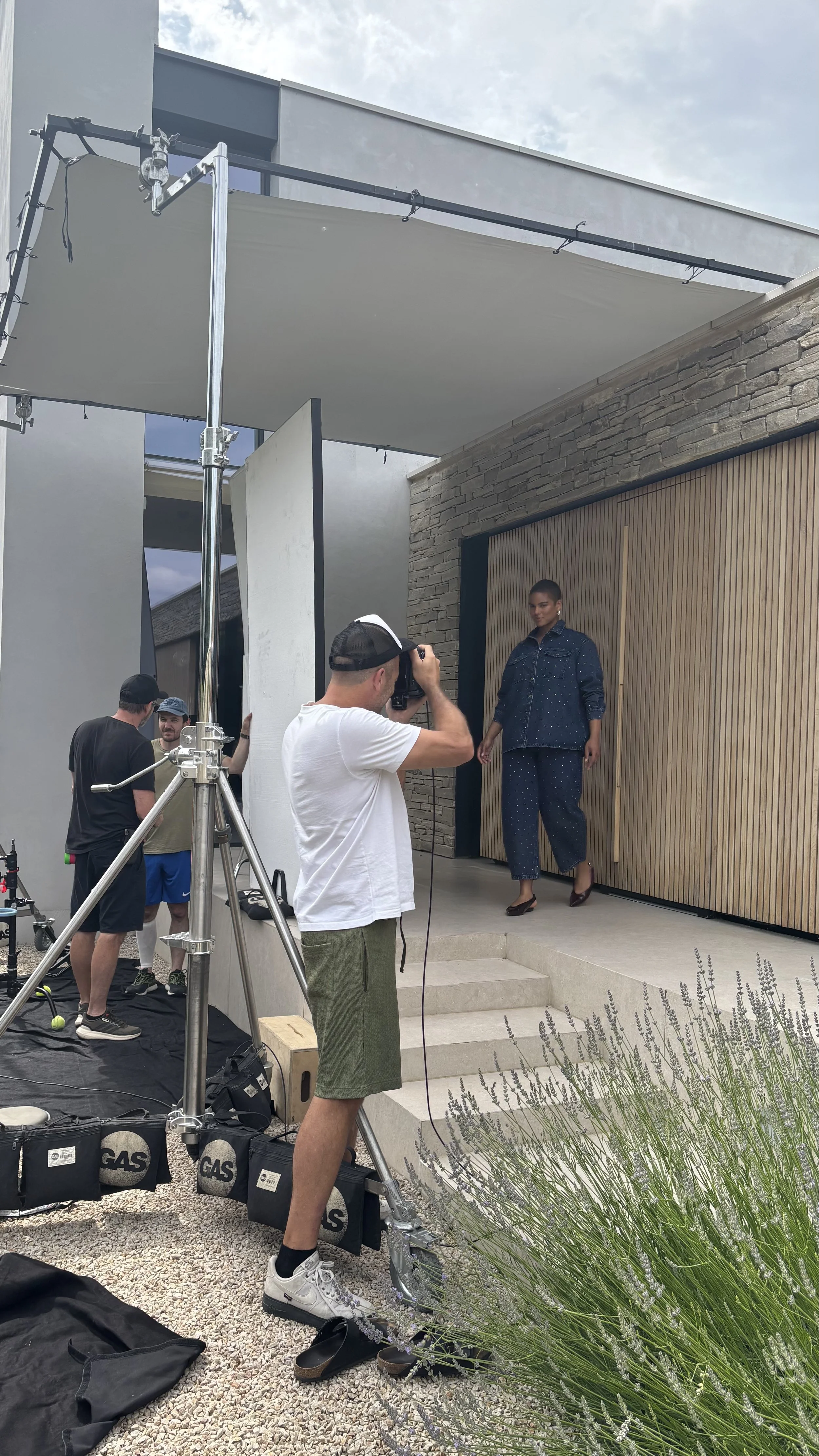 A photoshoot scene with a woman posing on steps outside a modern house, with a photographer taking her picture and others assisting, under a large overhead shade