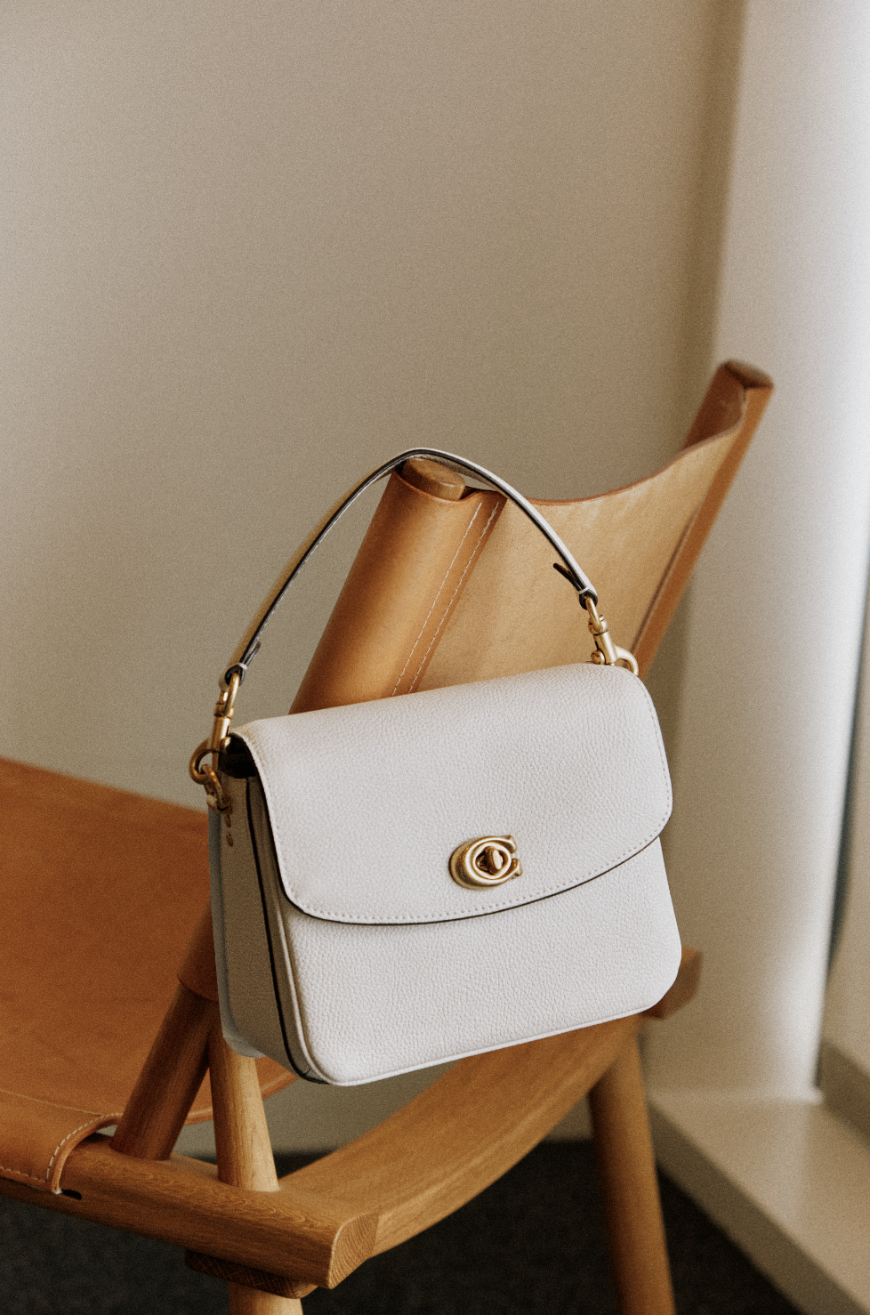A white handbag with a gold clasp hangs on the back of a wooden chair with a tan leather seat.