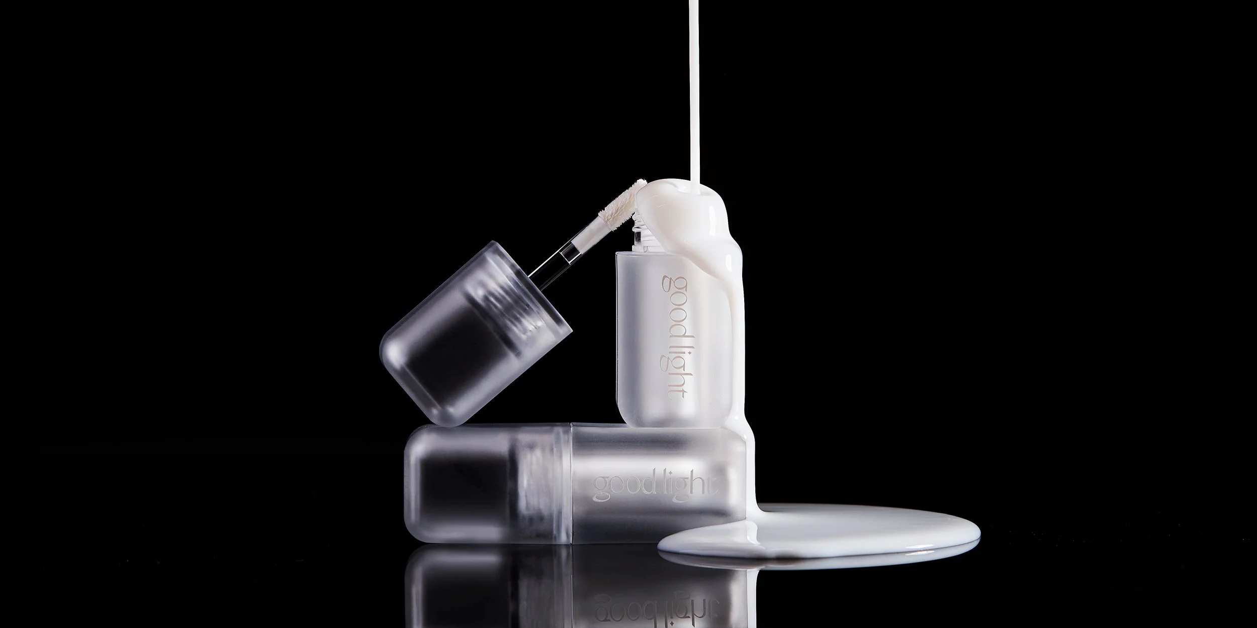 Three frosted nail polish bottles with the words "goodlight" on them, one pouring white paint or nail polish onto a reflective surface against a black background.