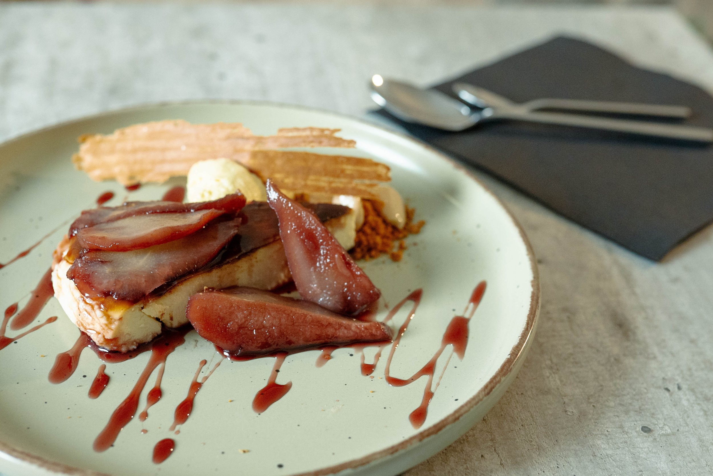 Postre de tarta de queso, frutas rojas, salsa de frutos rojos, y toques decorativos en un plato blanco sobre una mesa.
