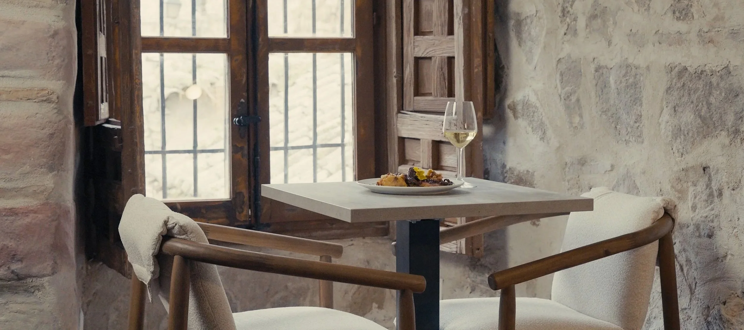 Mesa pequeña con plato de comida y una copa de vino blanco en un ambiente rústico con paredes de piedra y ventana de madera