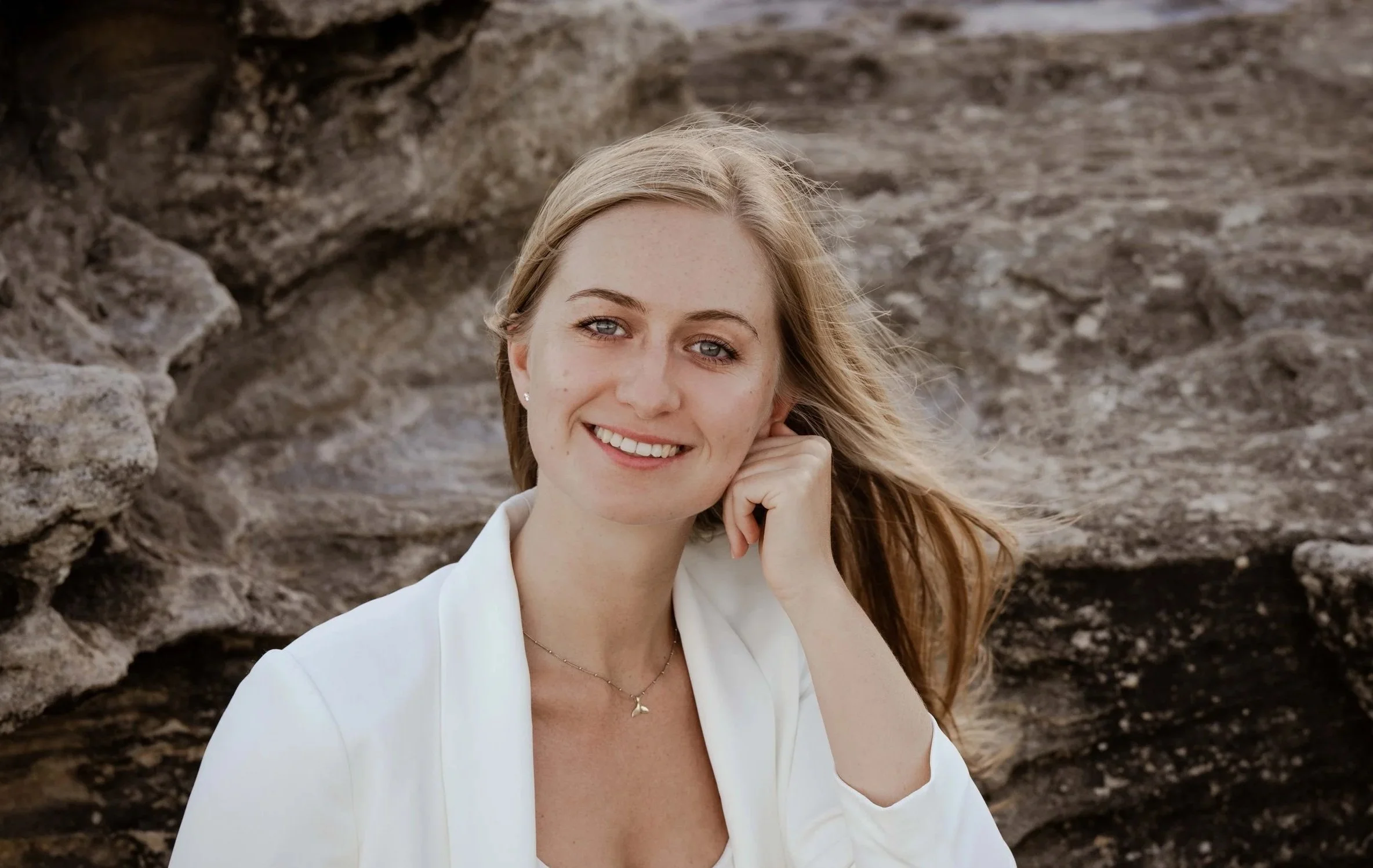 A woman with blonde hair, wearing a white blazer and a delicate necklace, seated outdoors against a rocky background, smiling at the camera.