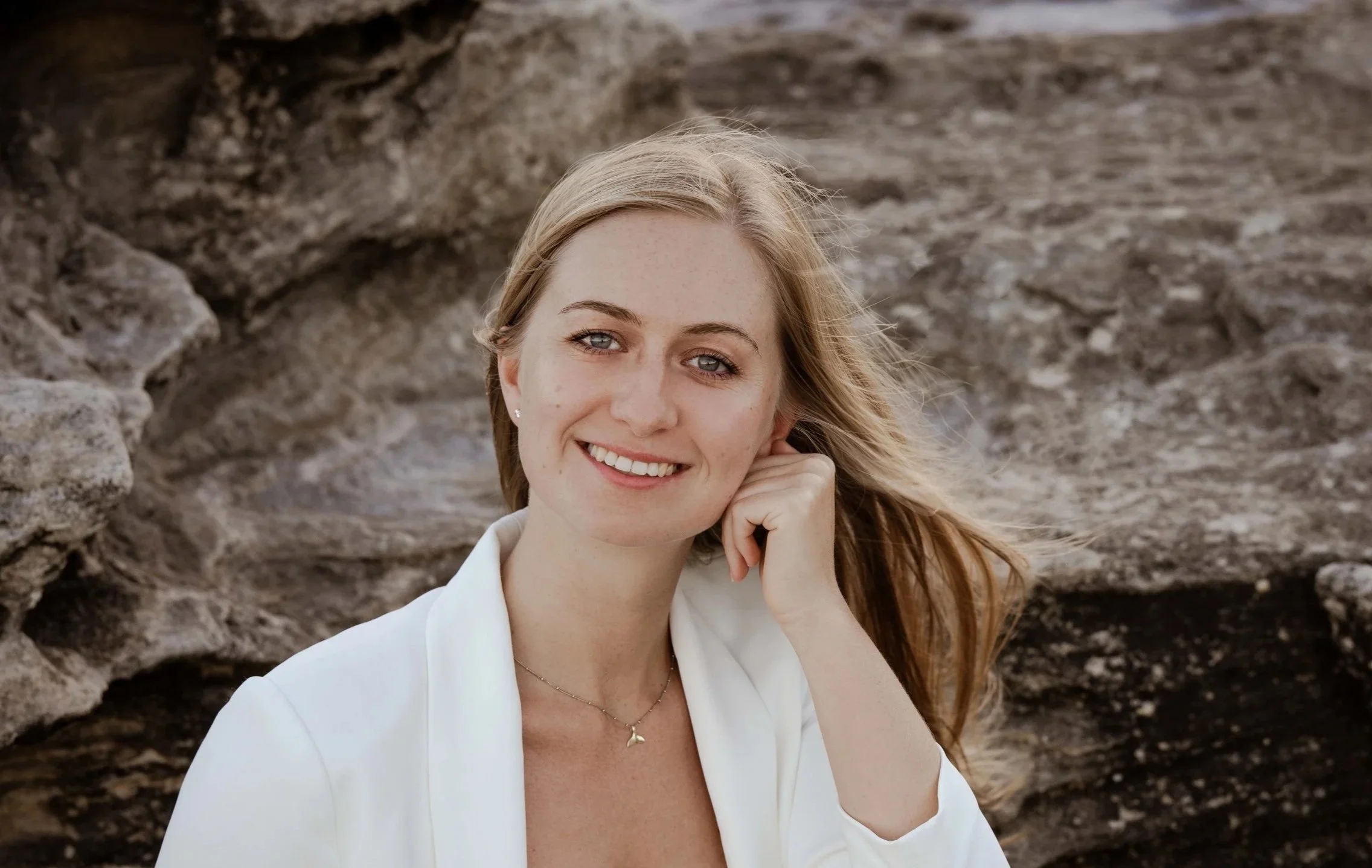 A smiling woman with long blonde hair, wearing a white blazer, standing outdoors with rocky background.