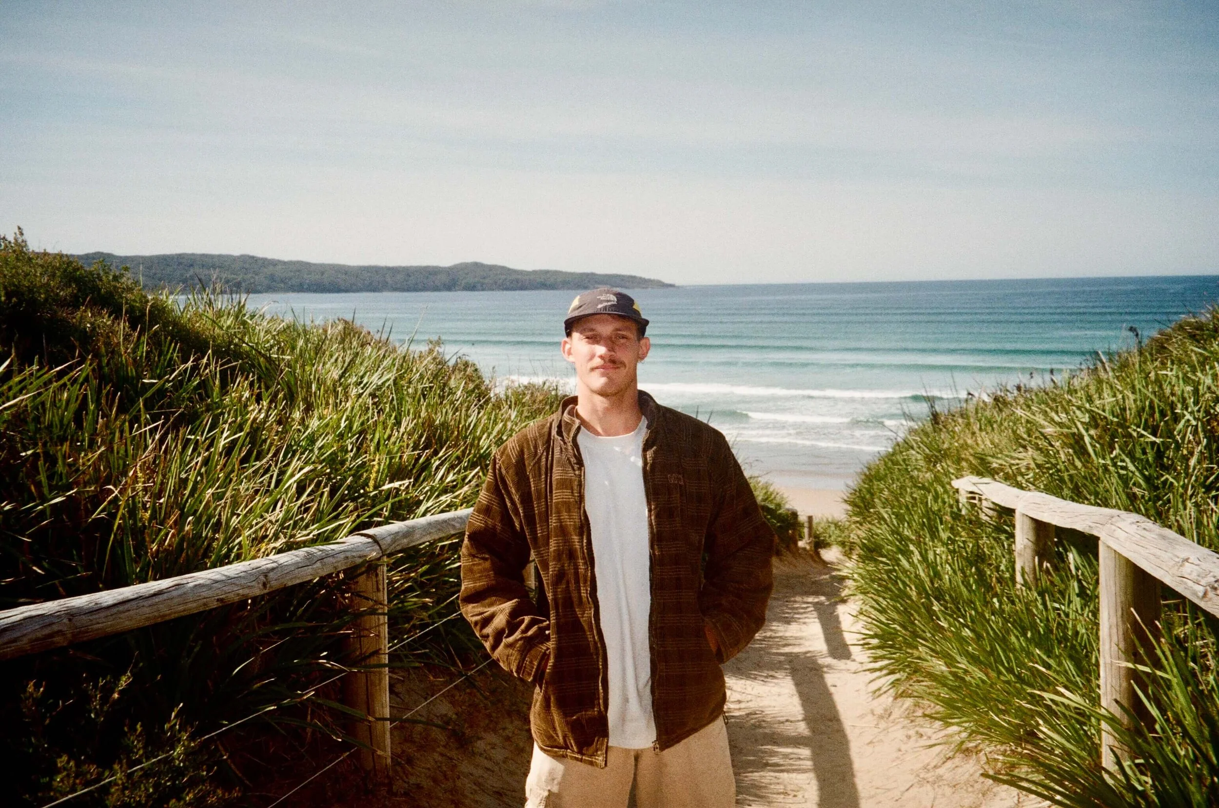 A young man stands on a sandy path leading to a beach, flanked by green bushes and wooden railings, with the ocean and sky in the background.