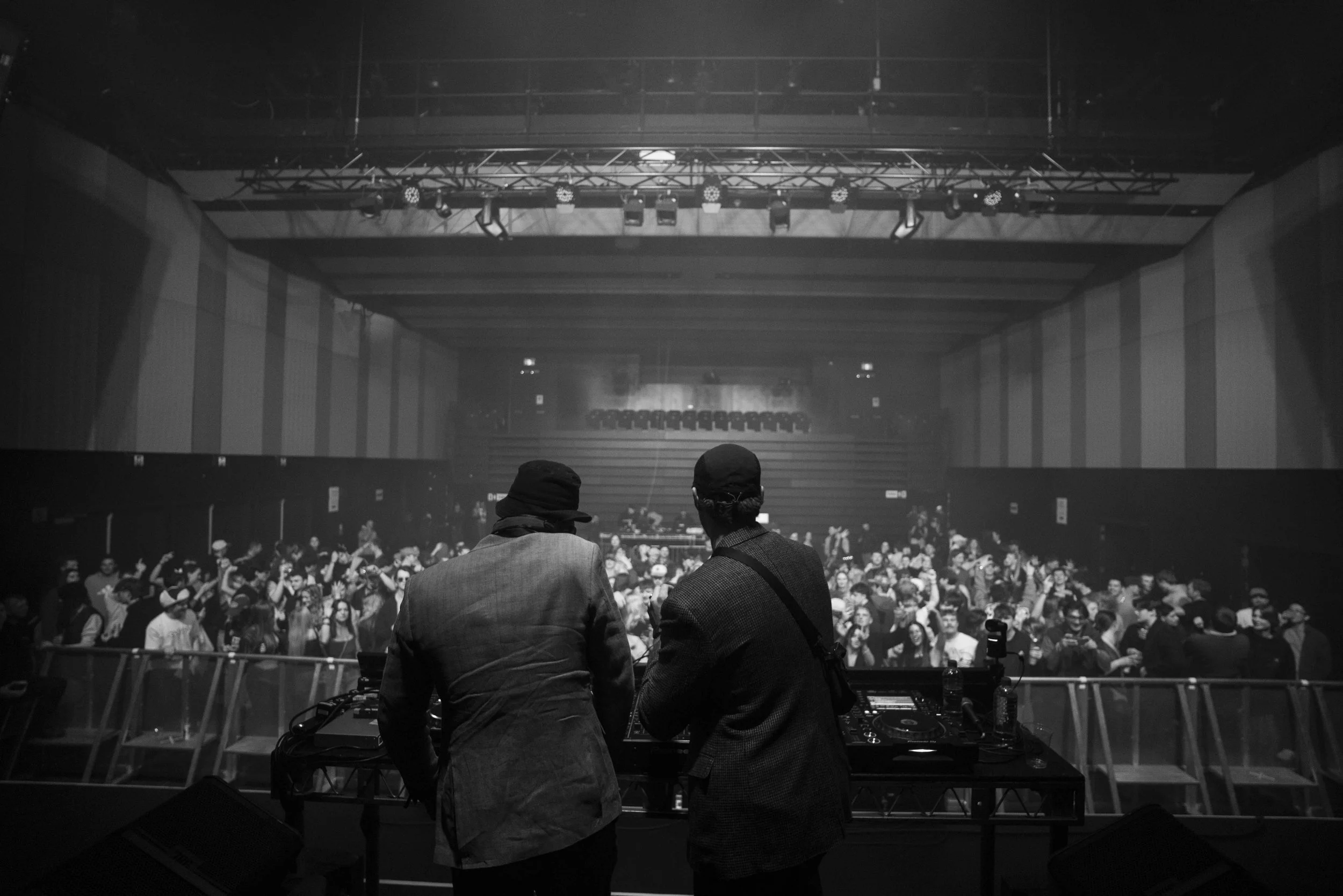 Two DJs in hats and suits stand behind turntables on stage in an indoor concert venue, facing a large crowd of people dancing and enjoying the music.