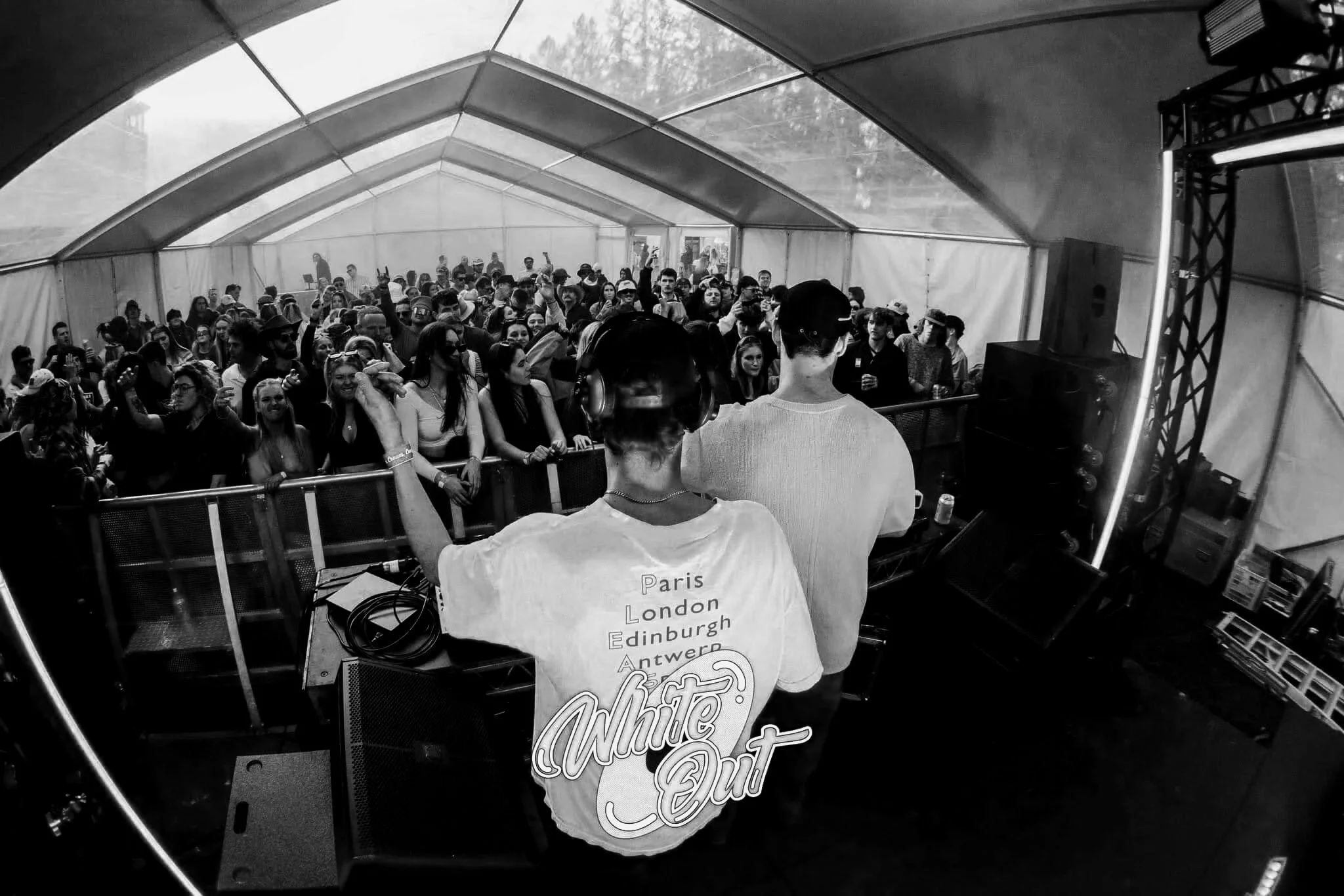 Two DJs perform at a music event in a tent, facing a large crowd of people.