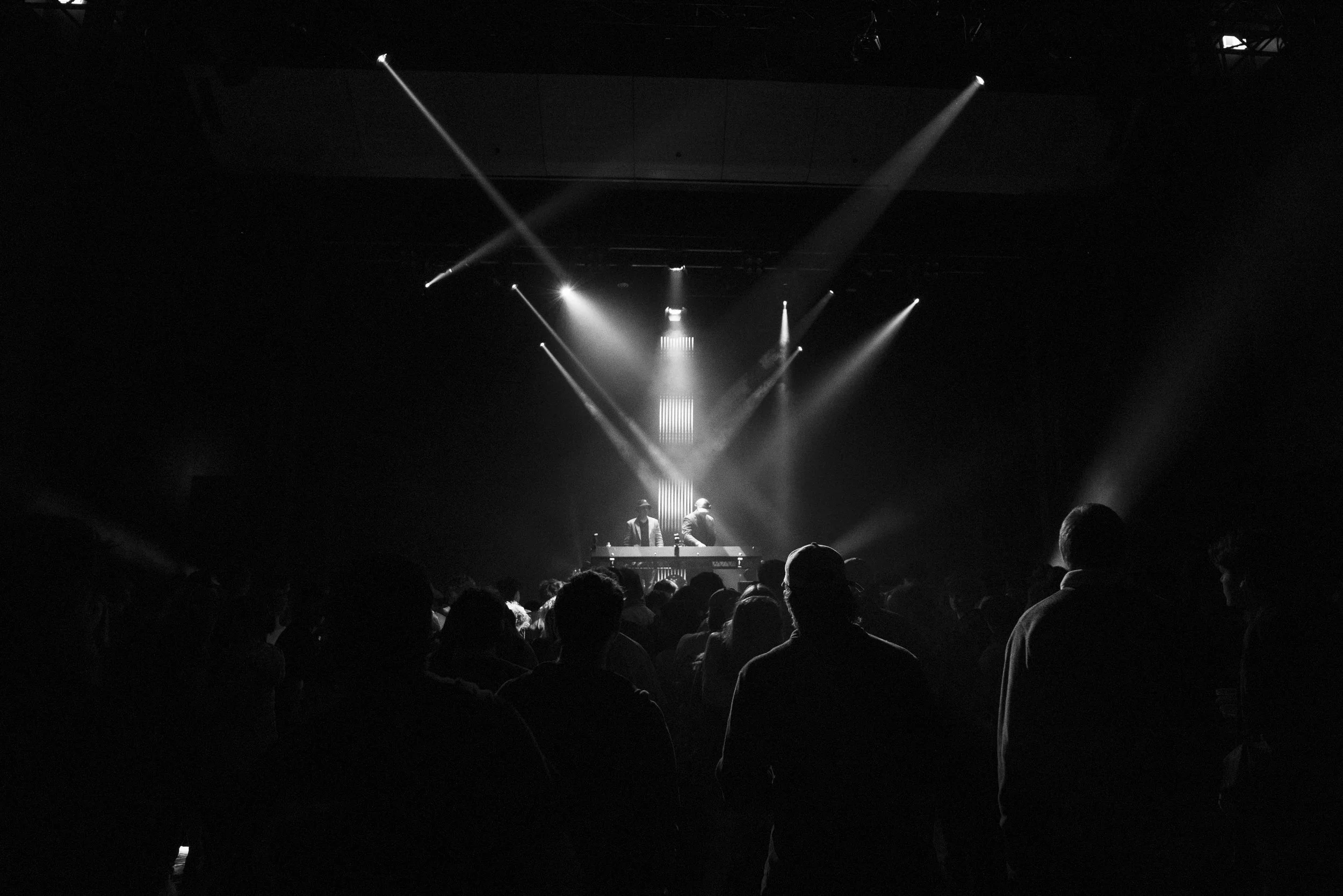 A black and white photo of a concert with a crowd facing a stage where two DJs are performing with lighting effects and smoke.