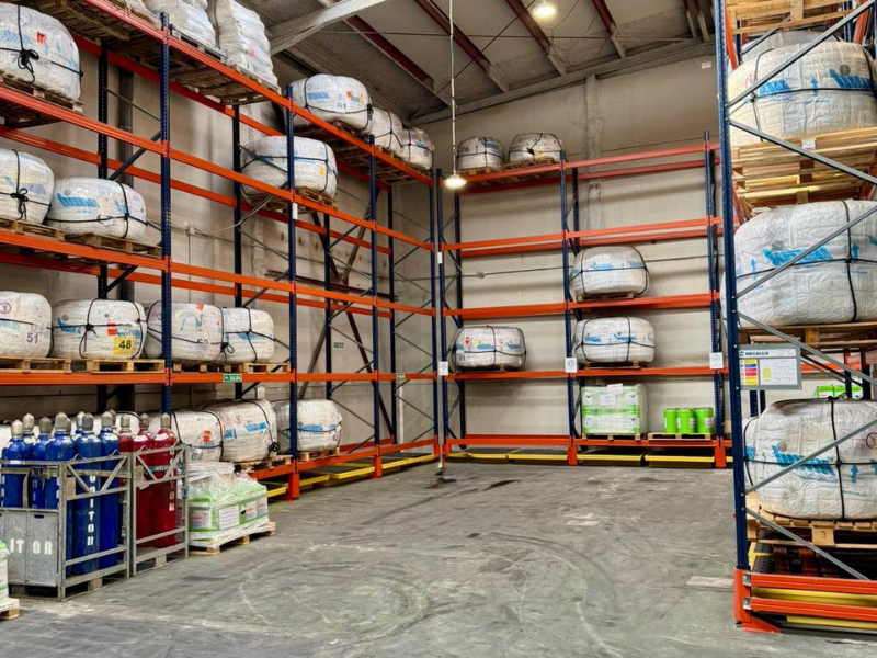 Empty industrial warehouse storage shelves with some wrapped packages and green watering cans, large open floor space, high ceiling with overhead lights, shelves made of orange and blue metal, and a rack holding gas cylinders.
