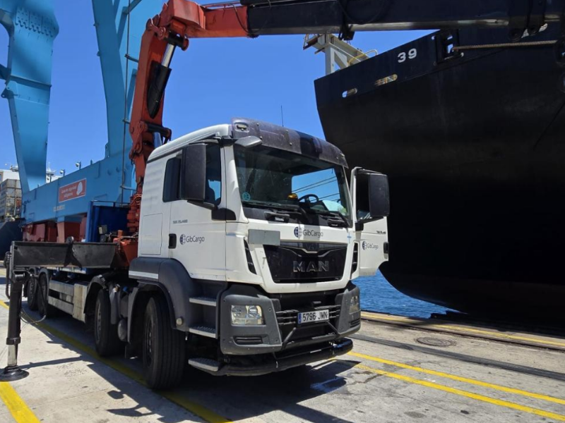 A cargo ship docked at a port with a large boat next to it. In the foreground, a white truck with a crane attachment is parked on the dock. The truck has a logo that reads 'Gbcargo'.