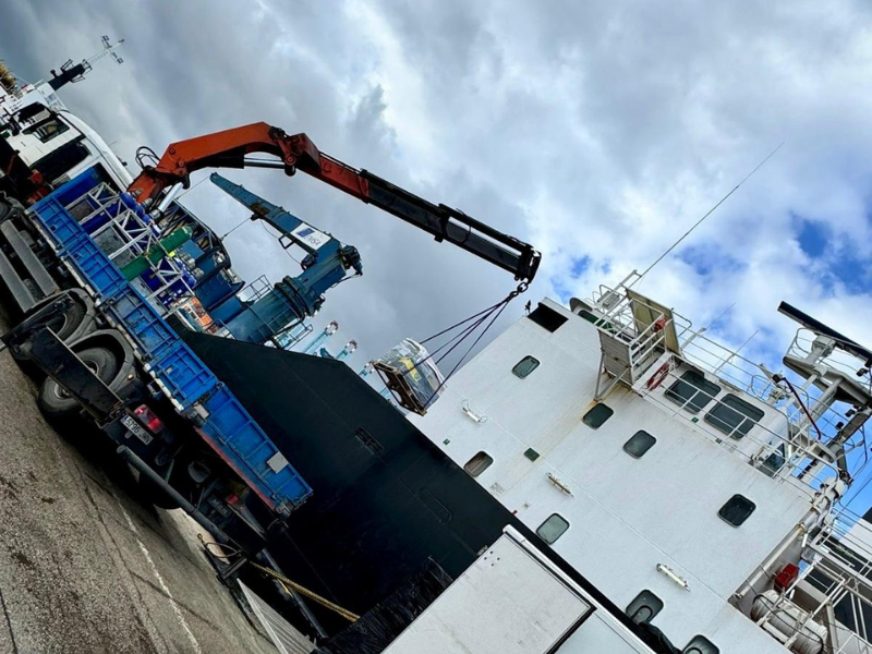 A cargo ship is being lifted by a crane at a dock, with a cloudy sky overhead.