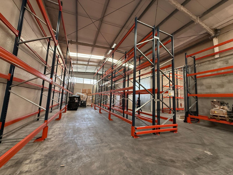 Empty industrial warehouse with orange and black metal storage shelves, some boxes and pallets at the far end.