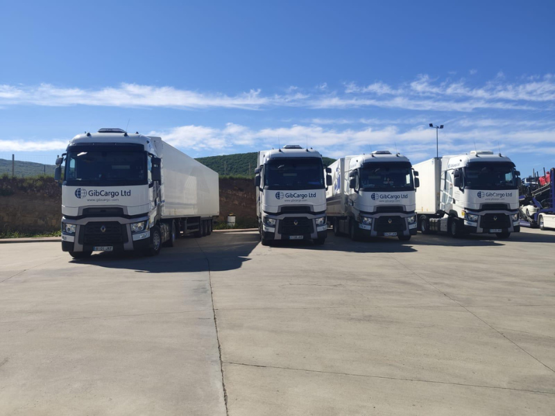Four white trucks with GibCargo Ltd logos parked on a concrete lot under a blue sky with scattered clouds.