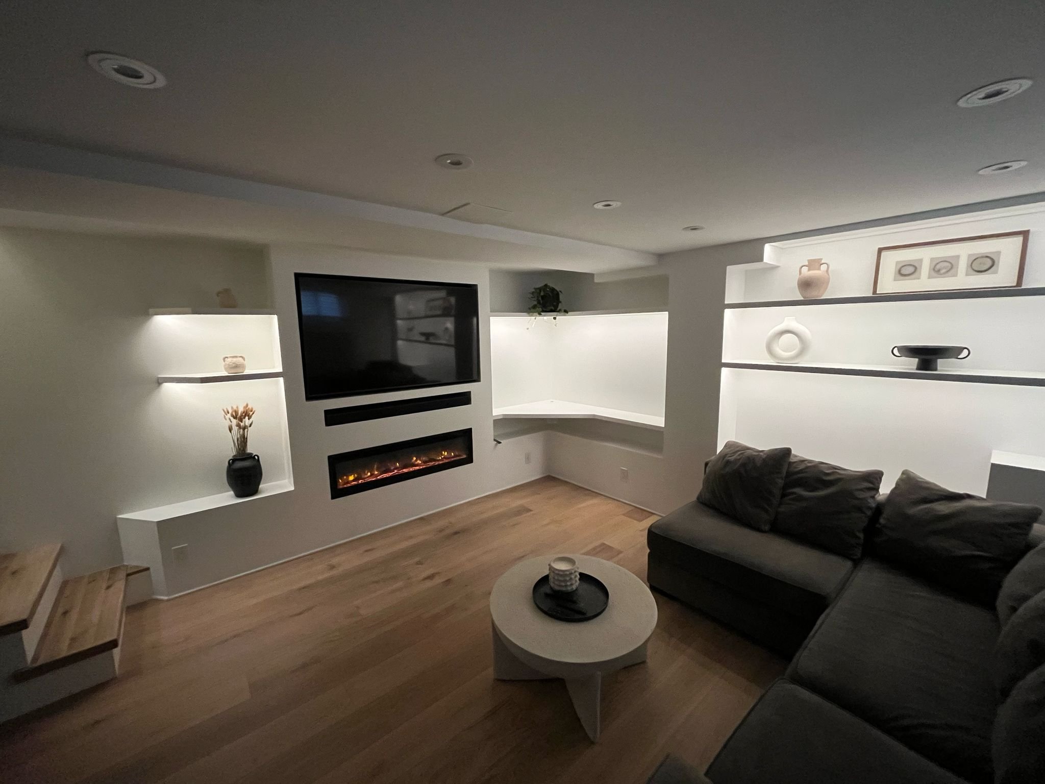 Modern living room with a mounted flat-screen TV, electric fireplace, wooden flooring, and built-in white wall shelves displaying vases and artwork. A dark sofa is visible along with a small round coffee table with decorative items.