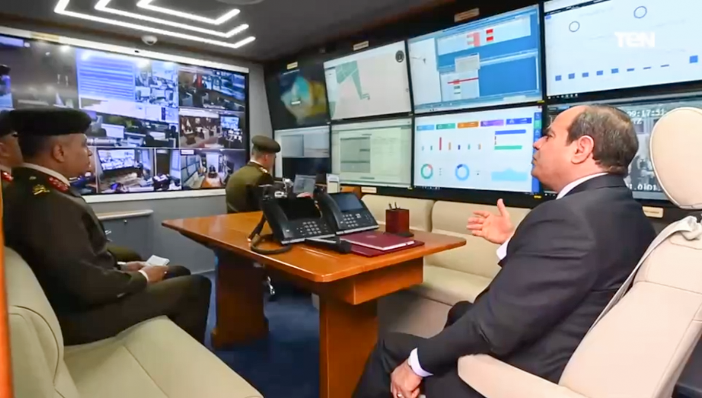 Man in a suit explaining data on large computer screens to military personnel in a control room.