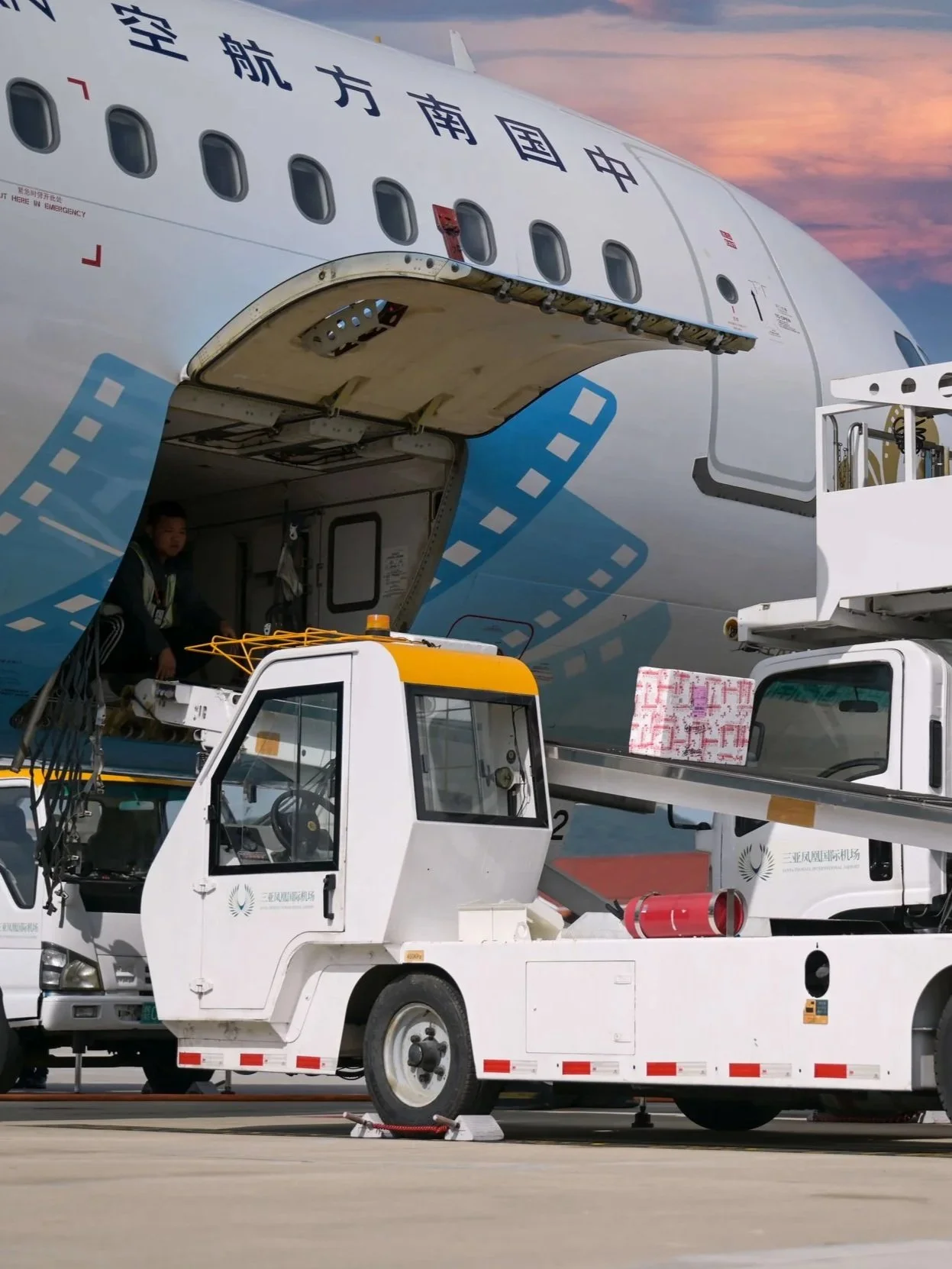 An airplane is parked at the gate with its door open for boarding. A ground service vehicle is positioned in front of the airplane, with a worker sitting on the platform. The scene is set during sunset, with a colorful sky in the background.