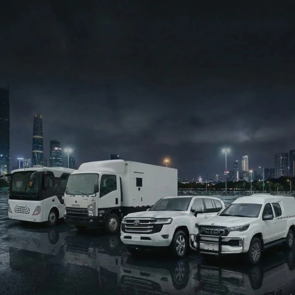 Four vehicles parked on a wet surface at night with a cityscape and dark clouds in the background. The vehicles include a white luxury RV, a white truck, and two white service vans.