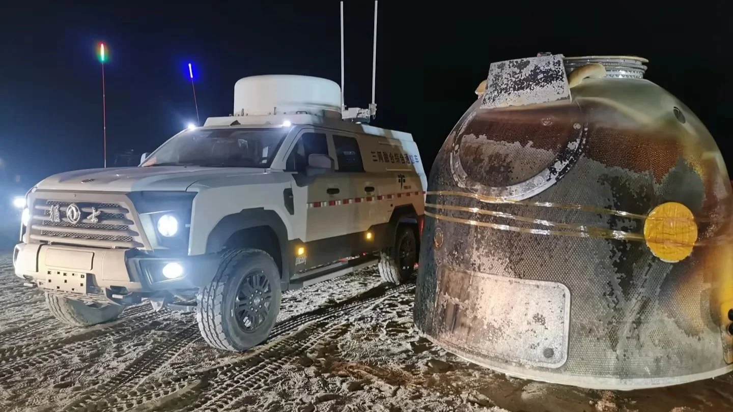 Nighttime scene with a military vehicle and a damaged spacecraft reentry capsule on the ground covered in snow or frost.