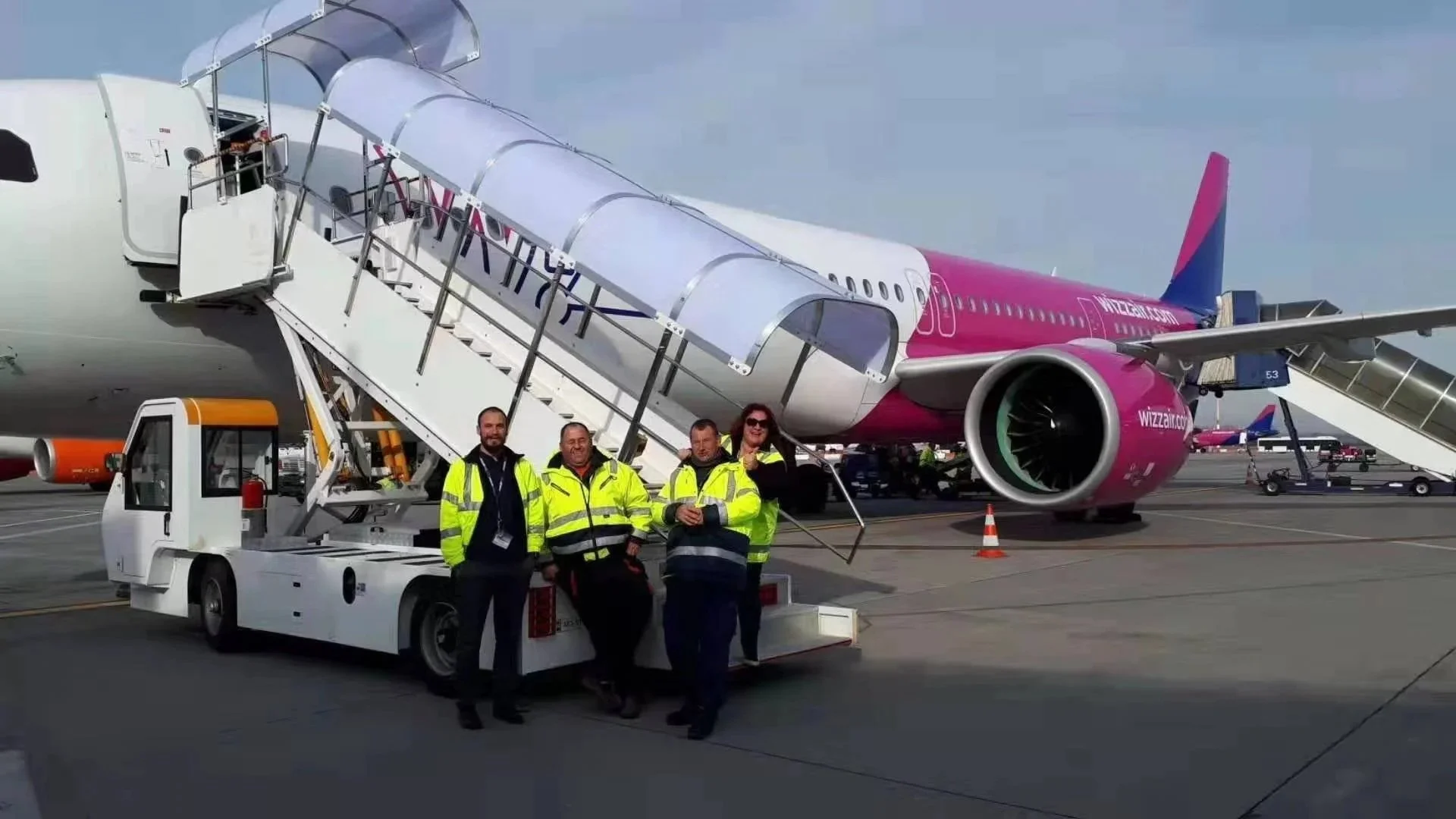 Four airline staff members in yellow safety jackets standing in front of a pink and white airplane at the airport, with stairs and a jet bridge connected to the aircraft.