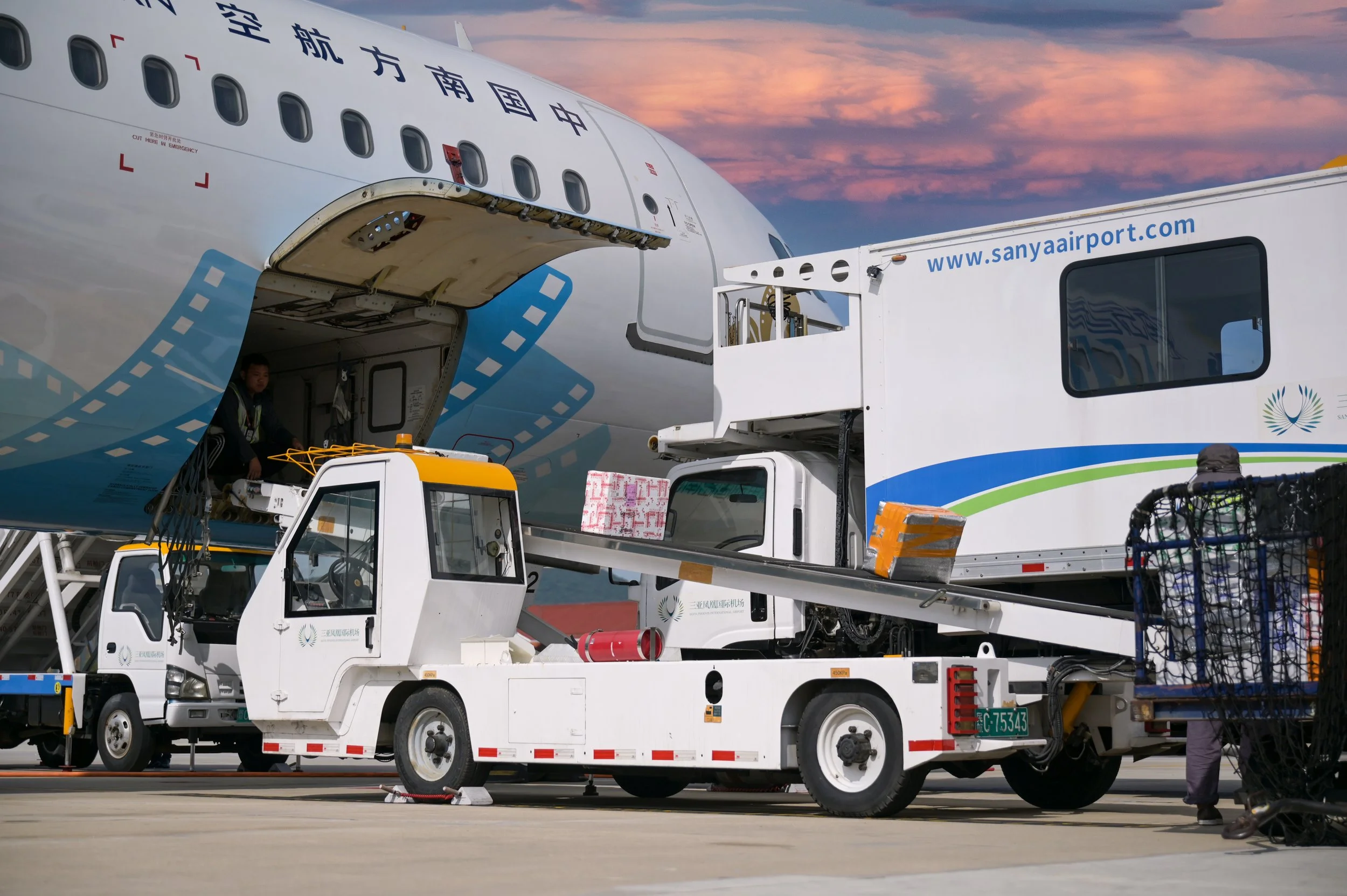 Airplane being loaded at Sanya Airport with ground crew and baggage carts, during sunset.