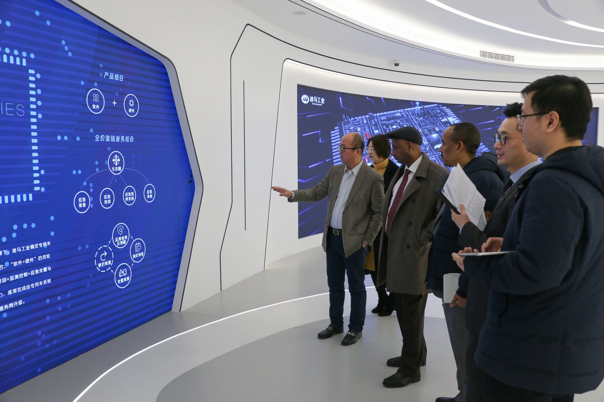 A group of six people observing and discussing a large digital screen displaying a blue interface with Chinese characters and icons in a modern, white-walled exhibition or conference space.