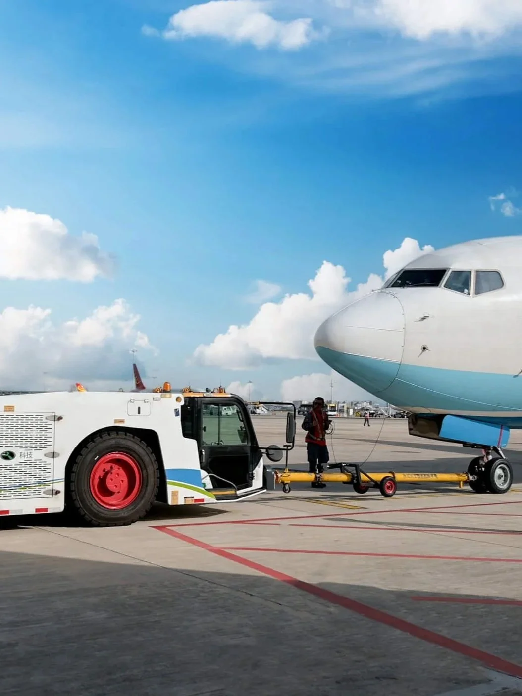An airplane on the tarmac being pushed by a ground vehicle. A person in a safety vest is nearby. The sky is partly cloudy.