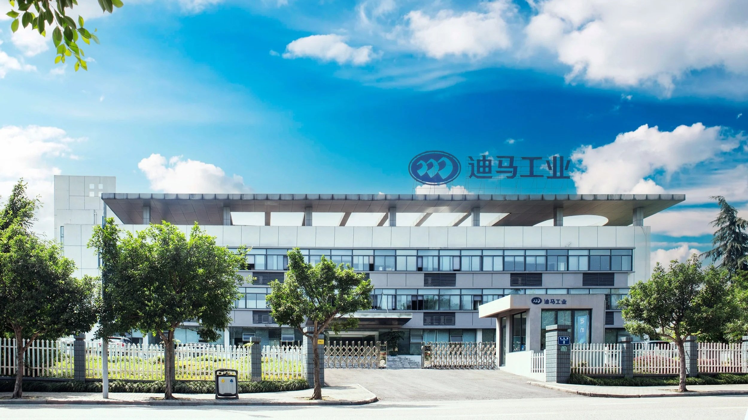 Modern industrial building with Chinese characters on the roof and a logo, surrounded by trees and a white fence, under a partly cloudy blue sky.