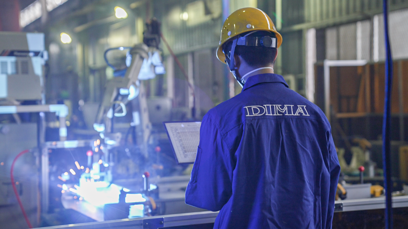 A worker in a factory wearing a yellow safety helmet, blue coveralls with a logo, and a face mask, looking at a control panel while industrial machinery operates nearby.