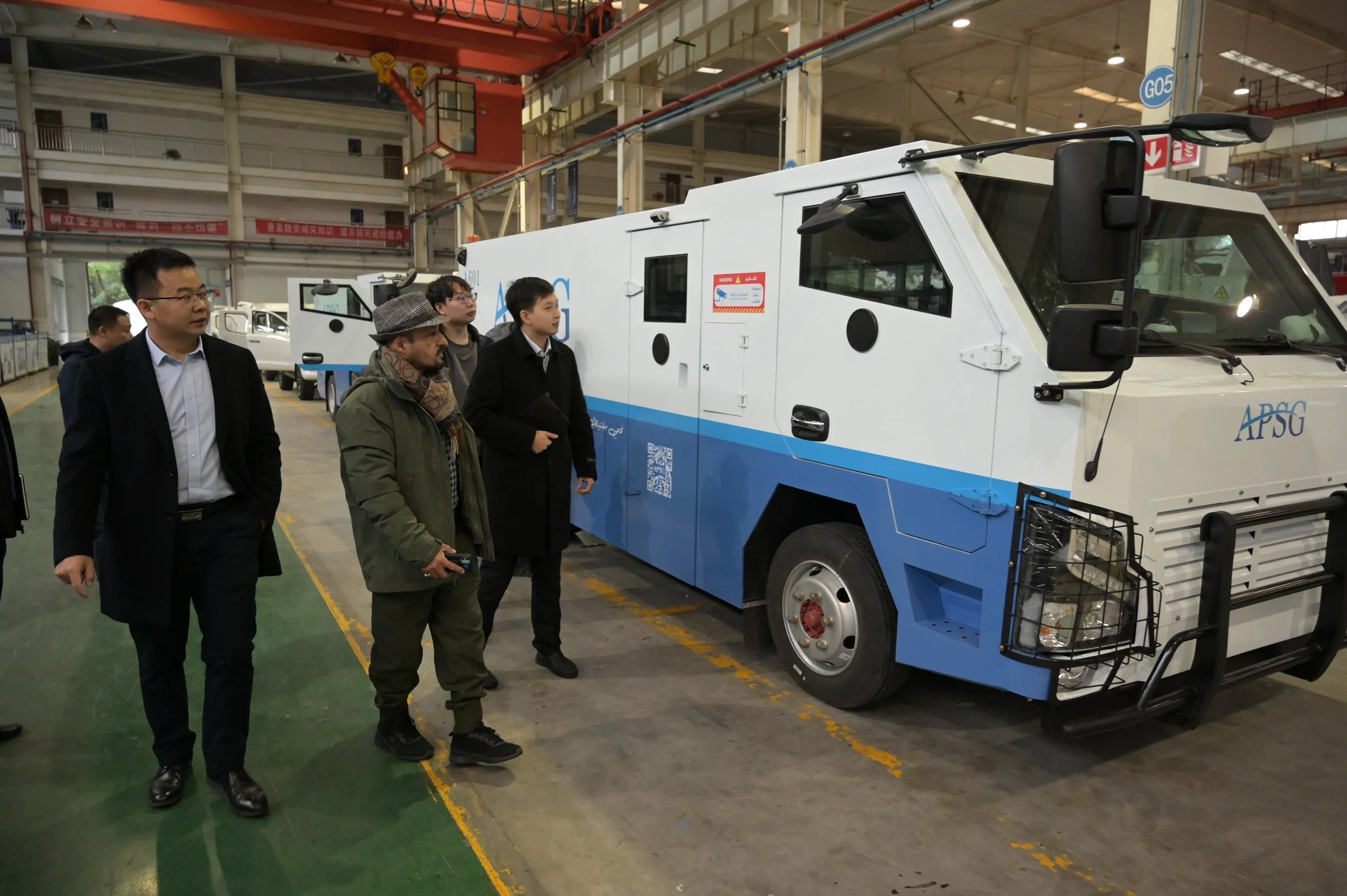 Group of men standing inside an industrial facility observing a large, white and blue vehicle, possibly a specialized truck or machinery, with multiple windows and ventilation openings.