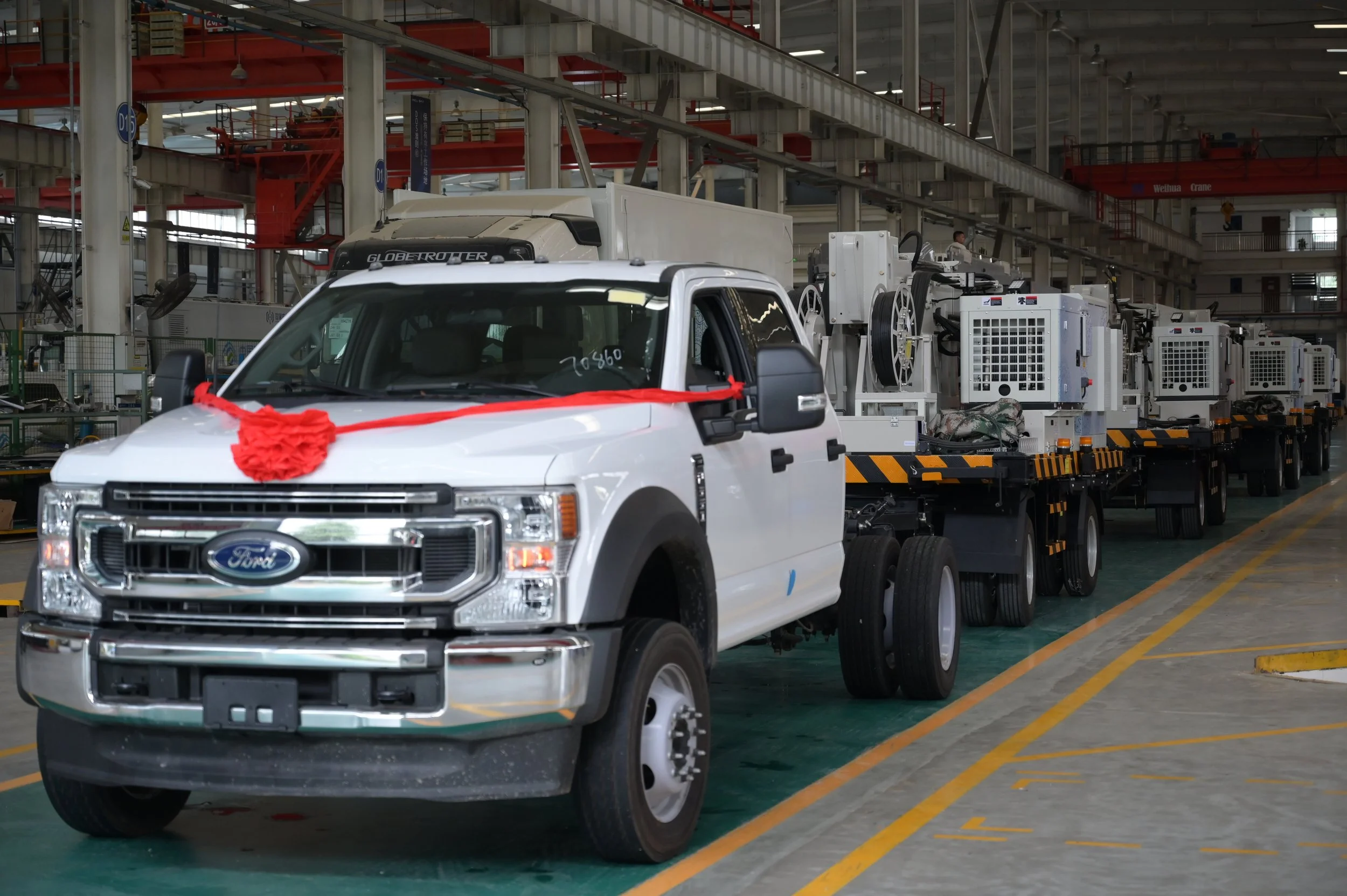 A white Ford flatbed truck decorated with a red ribbon, parked inside an industrial warehouse with machinery on the truck bed.