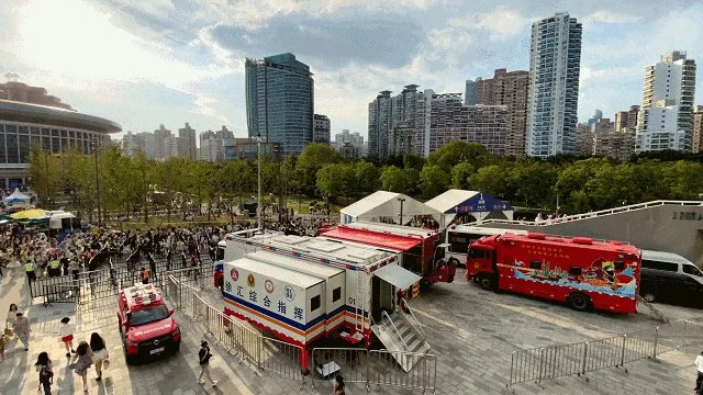 Crowd gathered outside a large white tent in an urban park with tall city buildings in the background. Several vehicles, including a red bus and a mobile command center, are parked nearby.