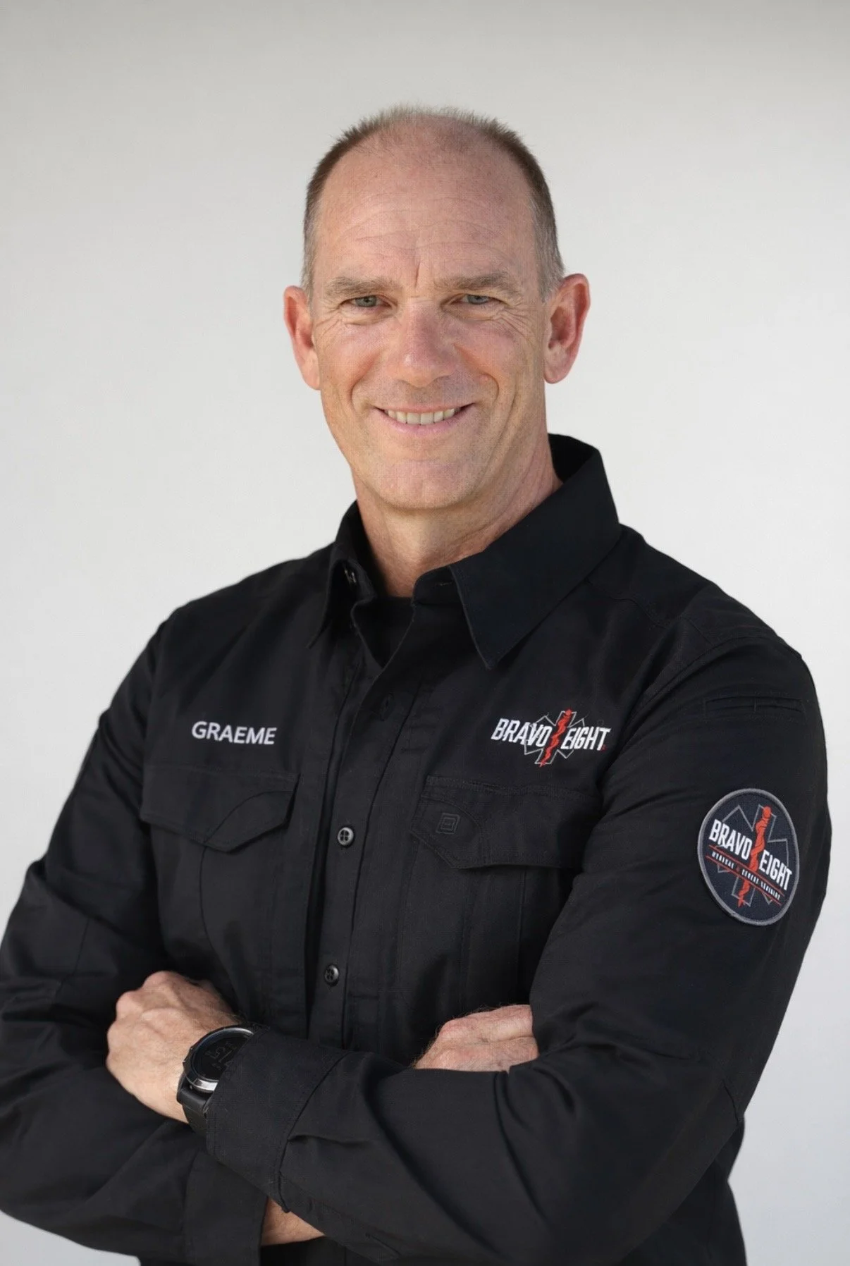 A man in a black shirt with 'GRAEME' embroidered on it, smiling with arms crossed, standing against a plain light background.