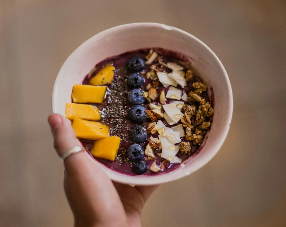 Hender som holder en açaí bowl med mango, blåbær, kokos og granola