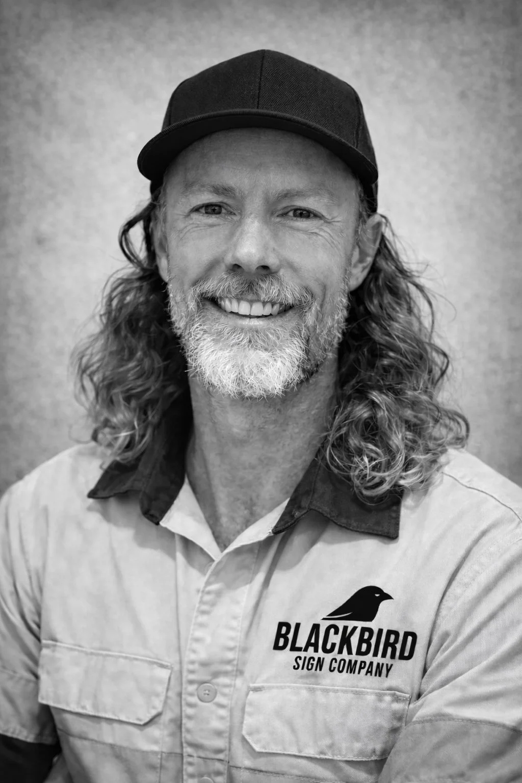 A smiling man with long wavy hair and a beard wearing a cap and a work shirt with the logo 'Blackbird Sign Company'
