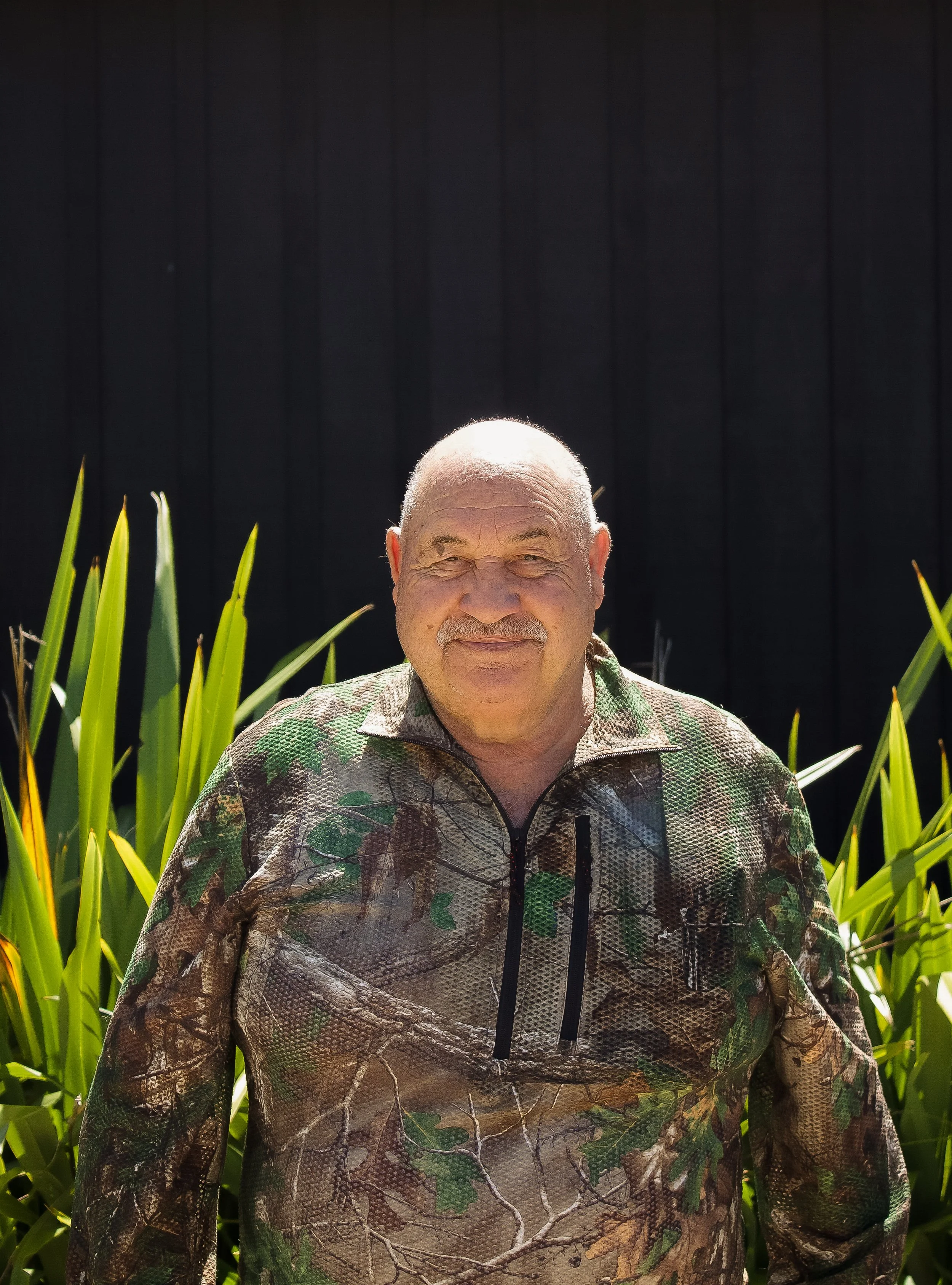 An elderly man with a bald head and a mustache smiling, wearing camouflage clothing, standing outdoors with tall green plants and a dark background.