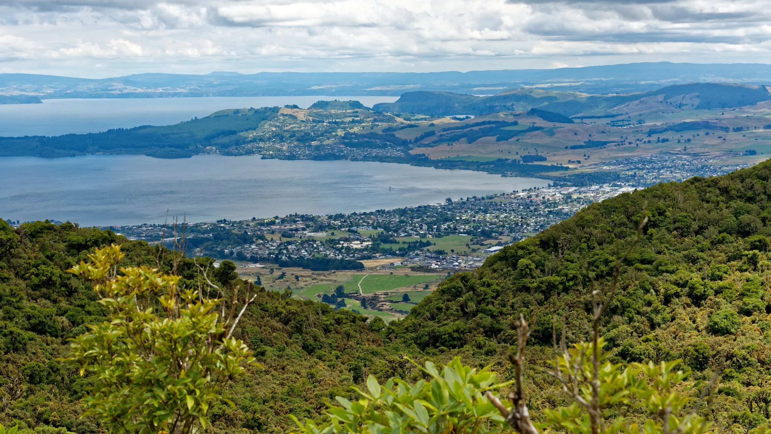 A scenic view of a large body of water with surrounding green hills, a city, and distant mountains under partly cloudy skies.