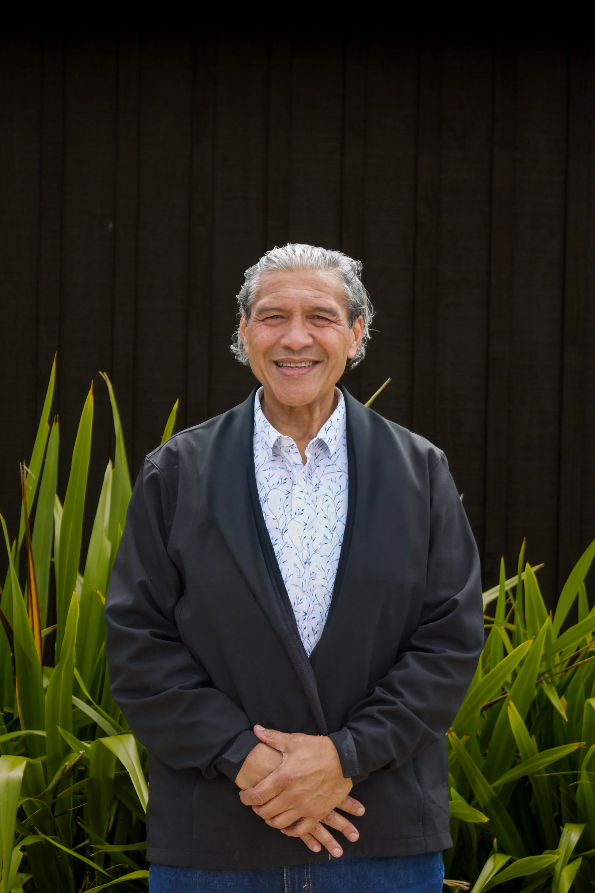 An elderly man with gray hair, smiling, standing outside in front of green plants and a dark wooden fence; wearing a black jacket and a white shirt with blue patterns.