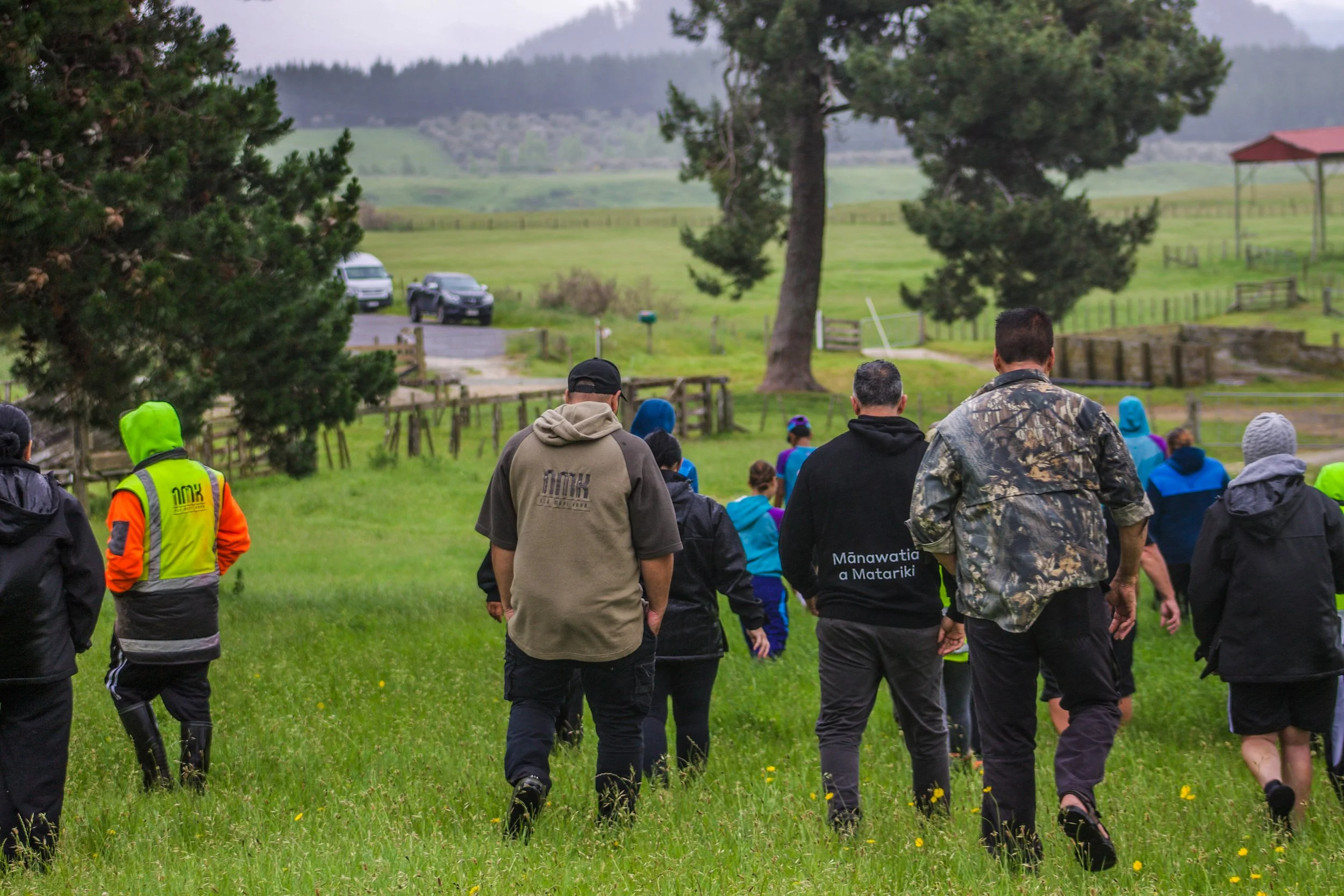 People walking in a grassy outdoor area with trees and a distant landscape, some wearing rain jackets and outdoor gear.